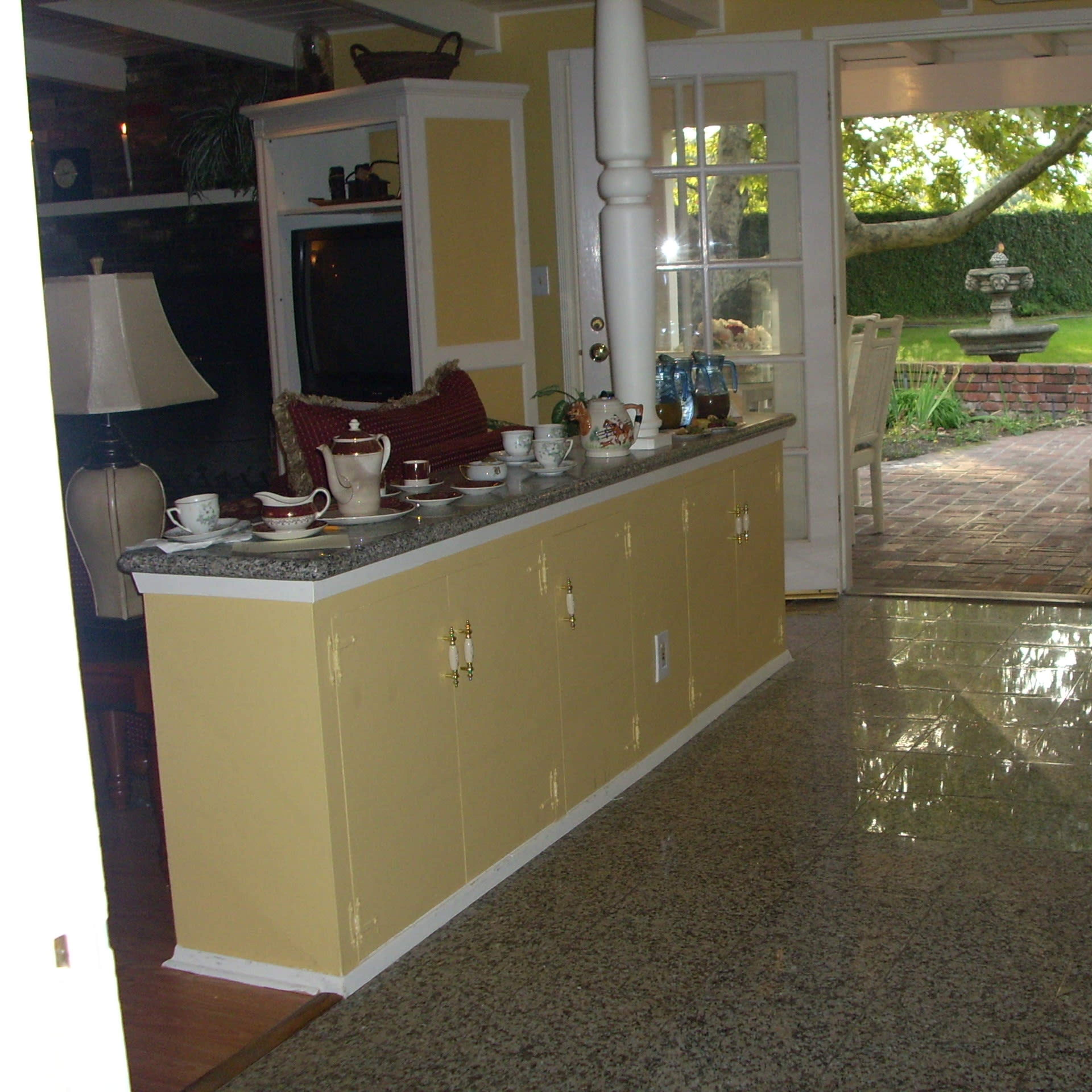 A spacious room with a yellow interior, featuring a granite countertop with tea sets and a view of a garden through an open doorway.