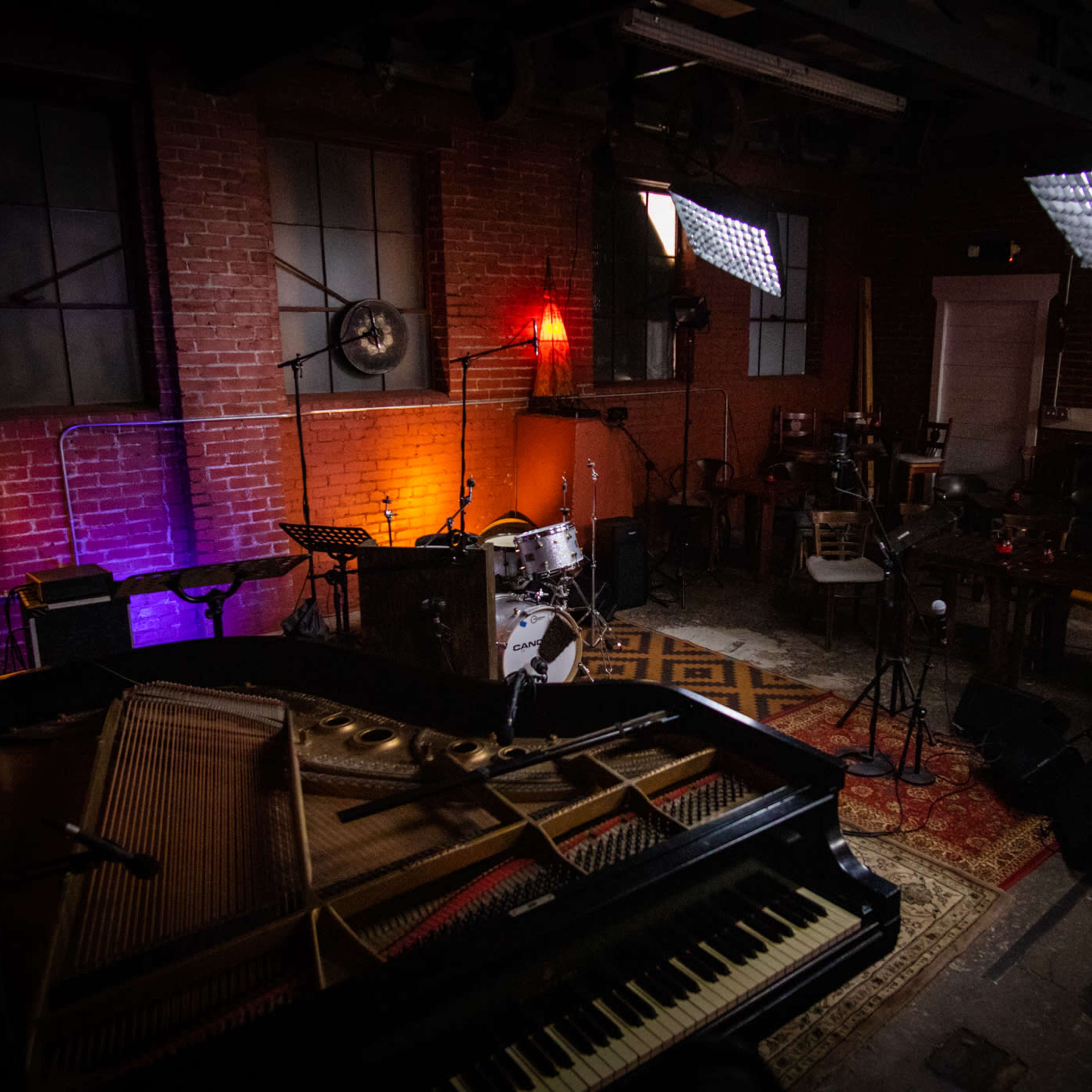 A dimly lit music venue with a grand piano in the foreground and various instruments set up, including a drum set and amplifiers against exposed brick walls.