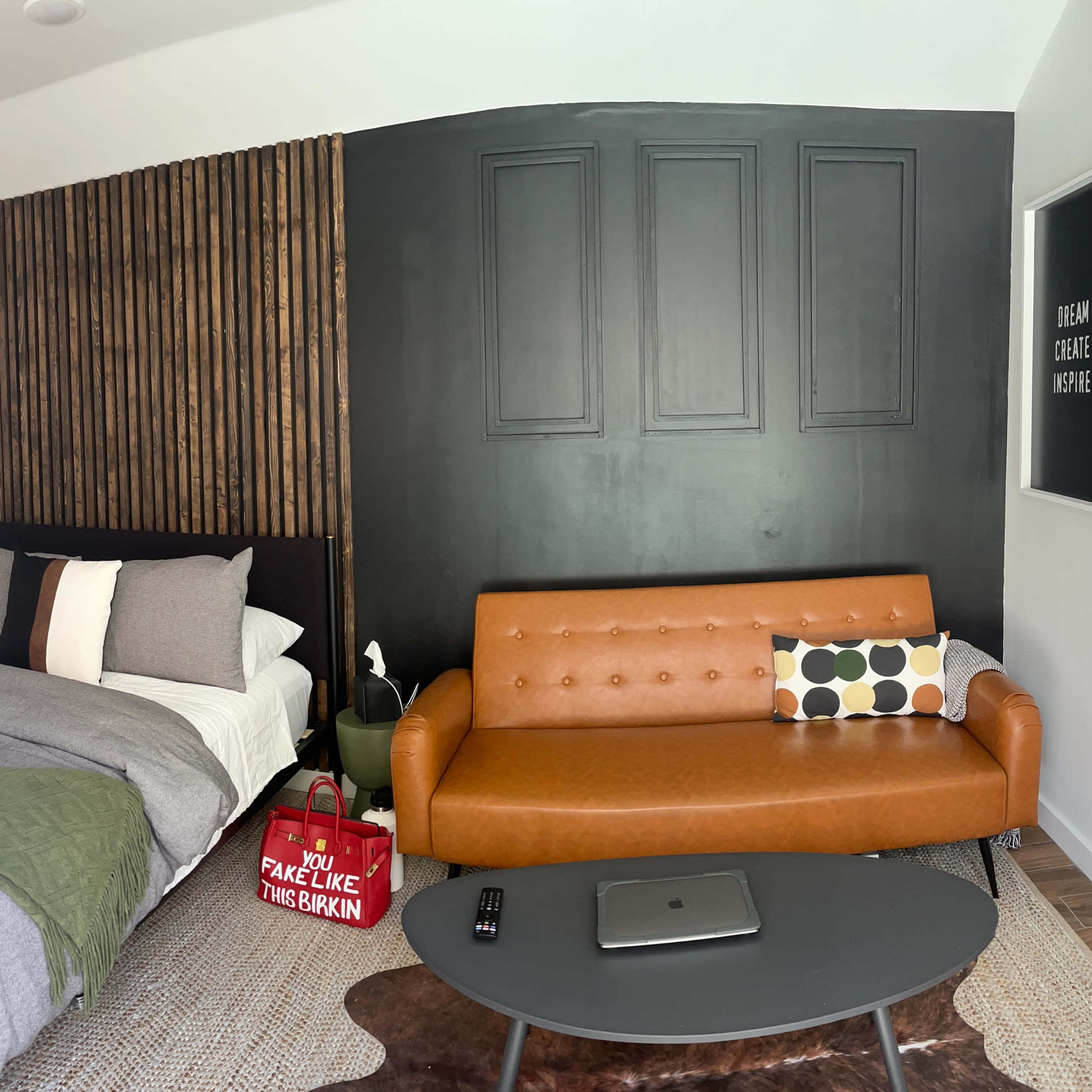 A small bedroom with a bed on one side and a brown couch with patterned cushions on the other, featuring a wall with wooden paneling and a dark painted section.