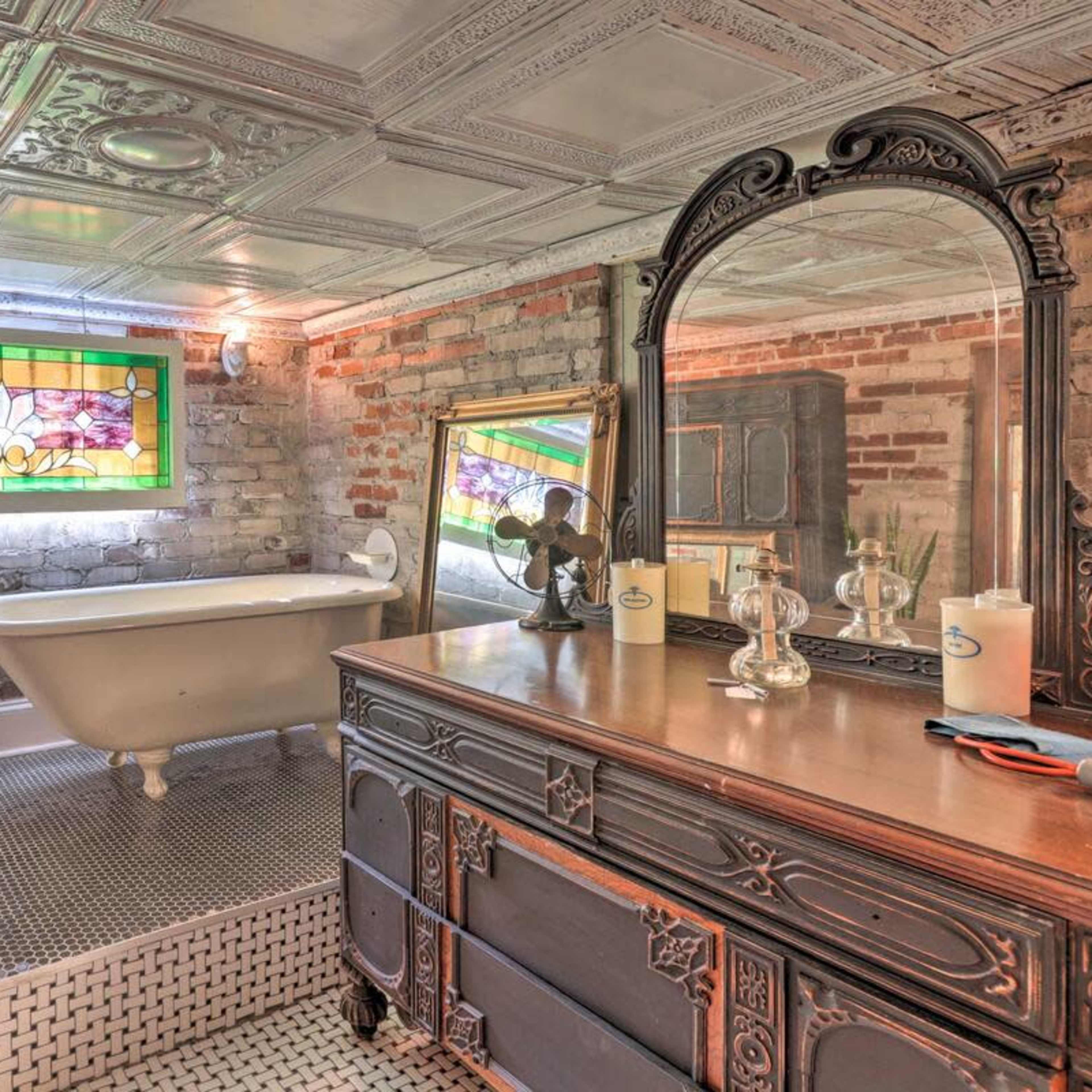 A vintage bathroom featuring a freestanding bathtub, brick walls, a stained glass window, and ornate wooden furniture.