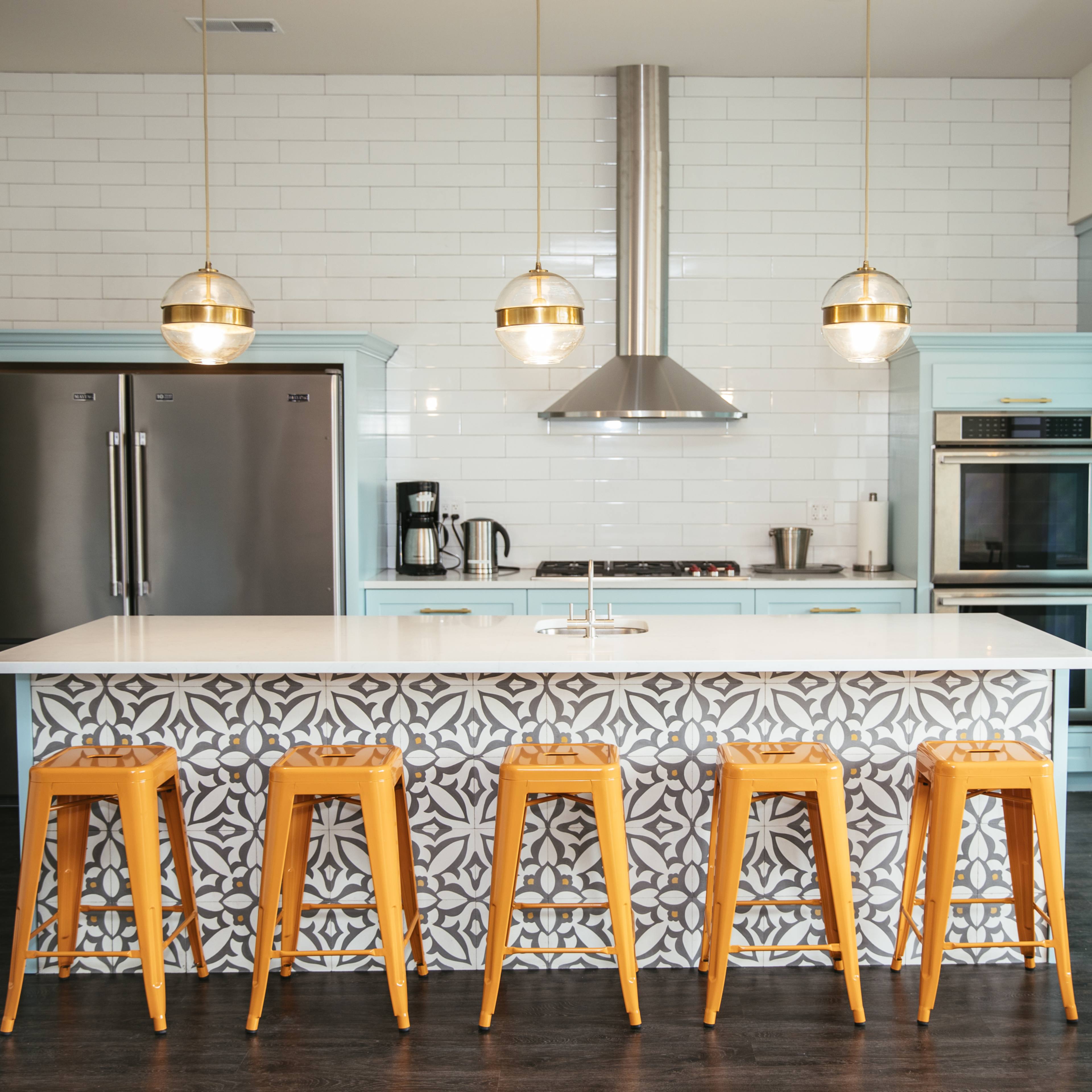 A modern kitchen features a large island with a patterned front, four yellow stools, stainless steel appliances, and pendant lights above.