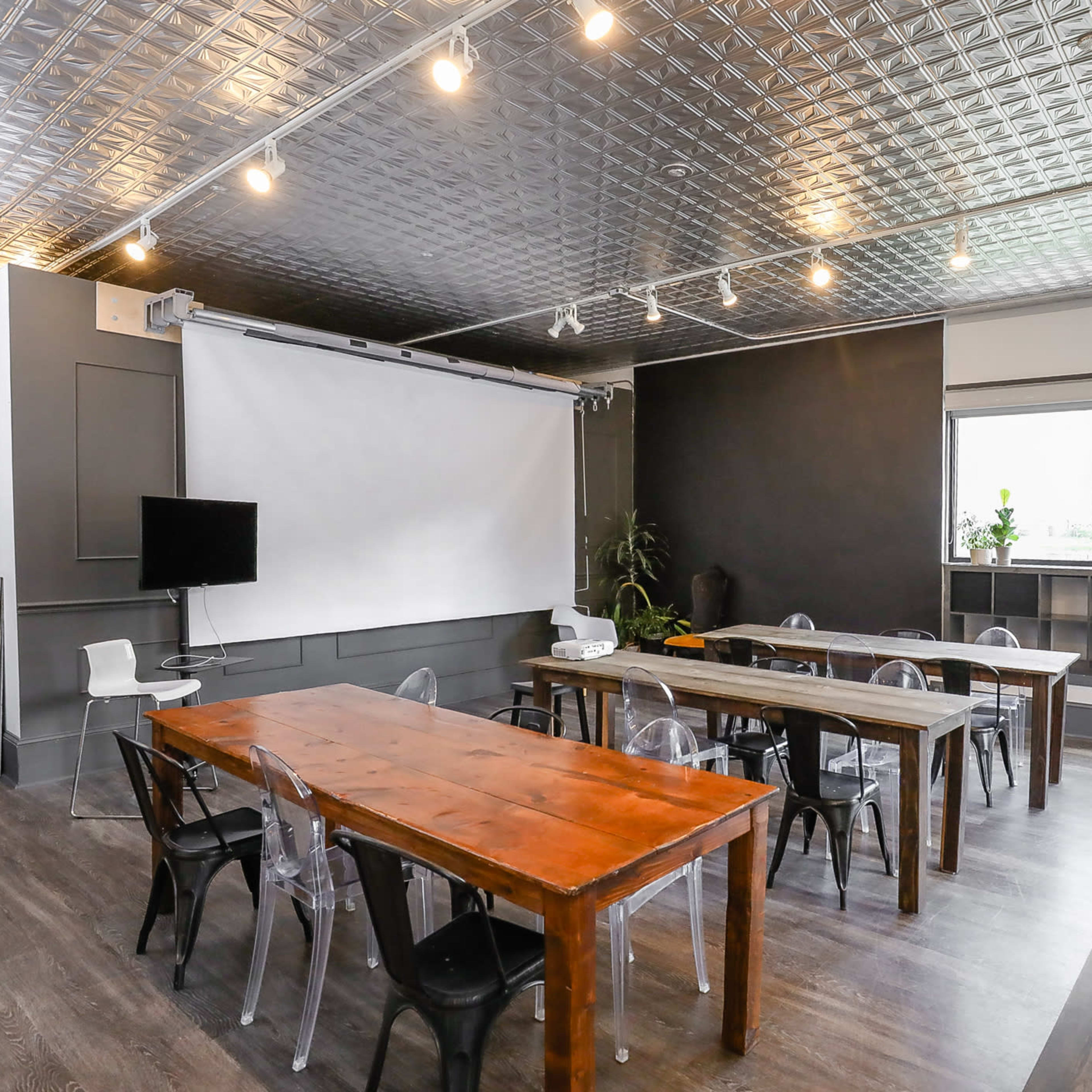 A modern meeting room with two long wooden tables, transparent chairs, a projector screen, and a TV on the wall.