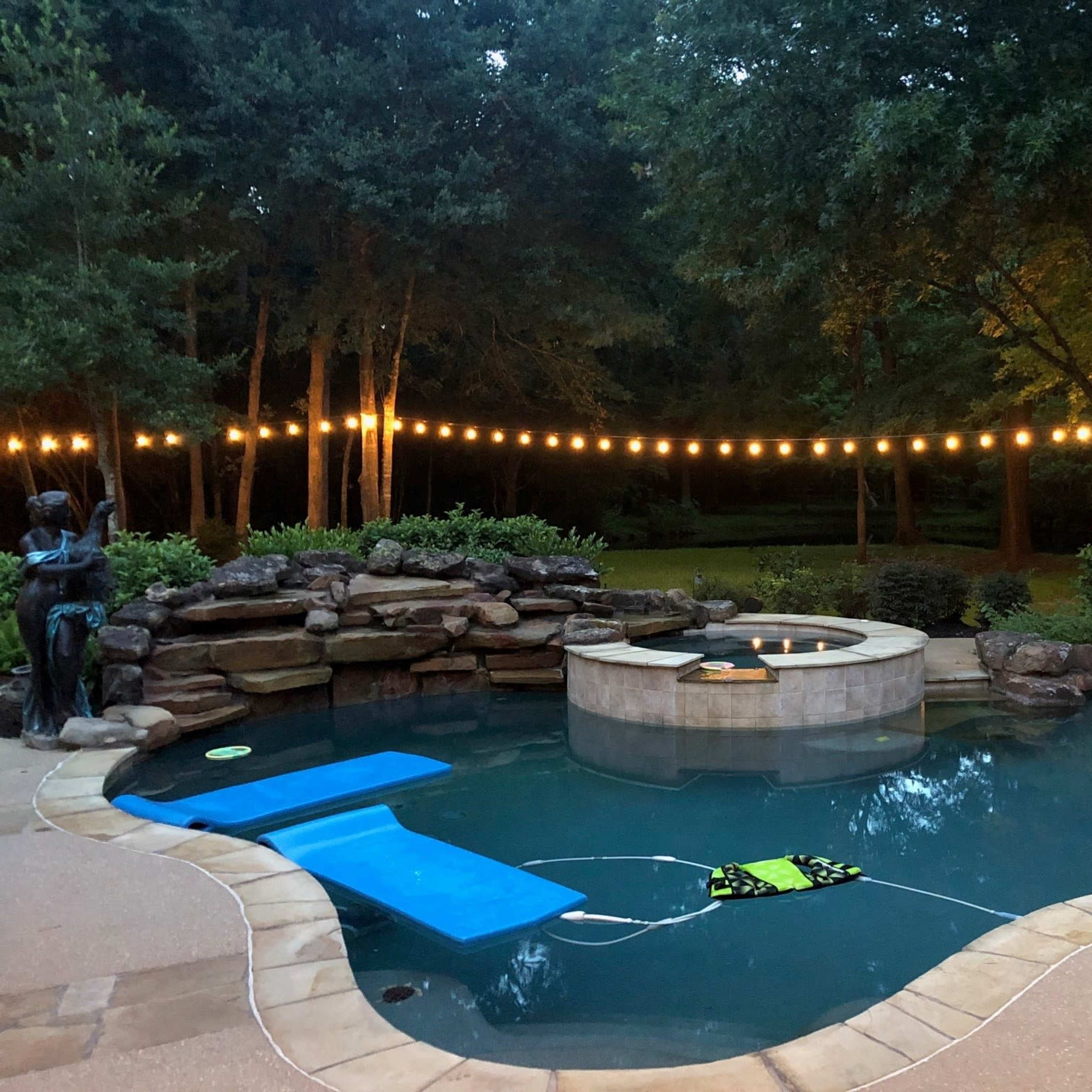 A backyard pool with a raised hot tub, surrounded by a stone wall and illuminated by string lights overhead.