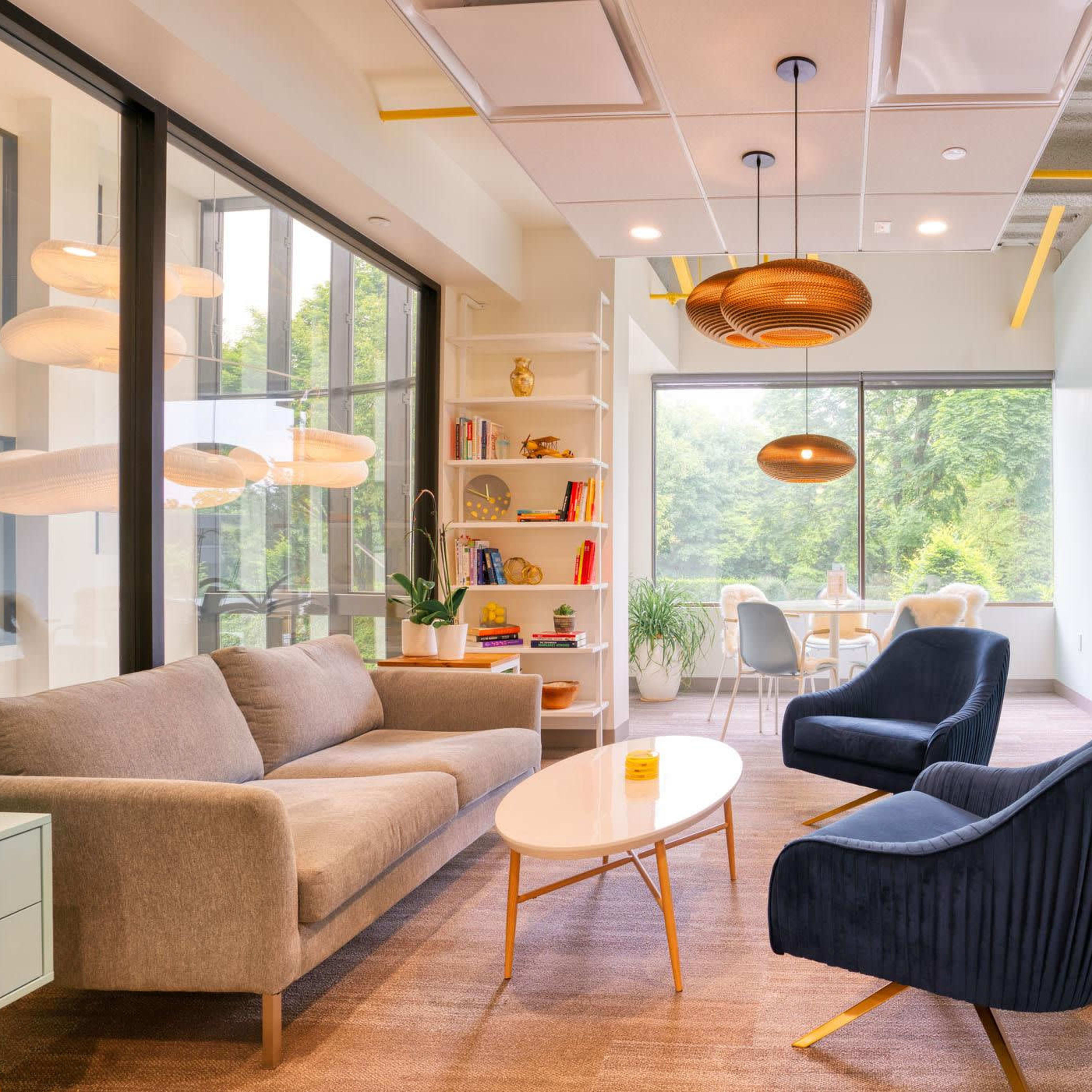 A modern office lounge area featuring a gray sofa, two blue armchairs, a round coffee table, and a large window with a view of greenery.