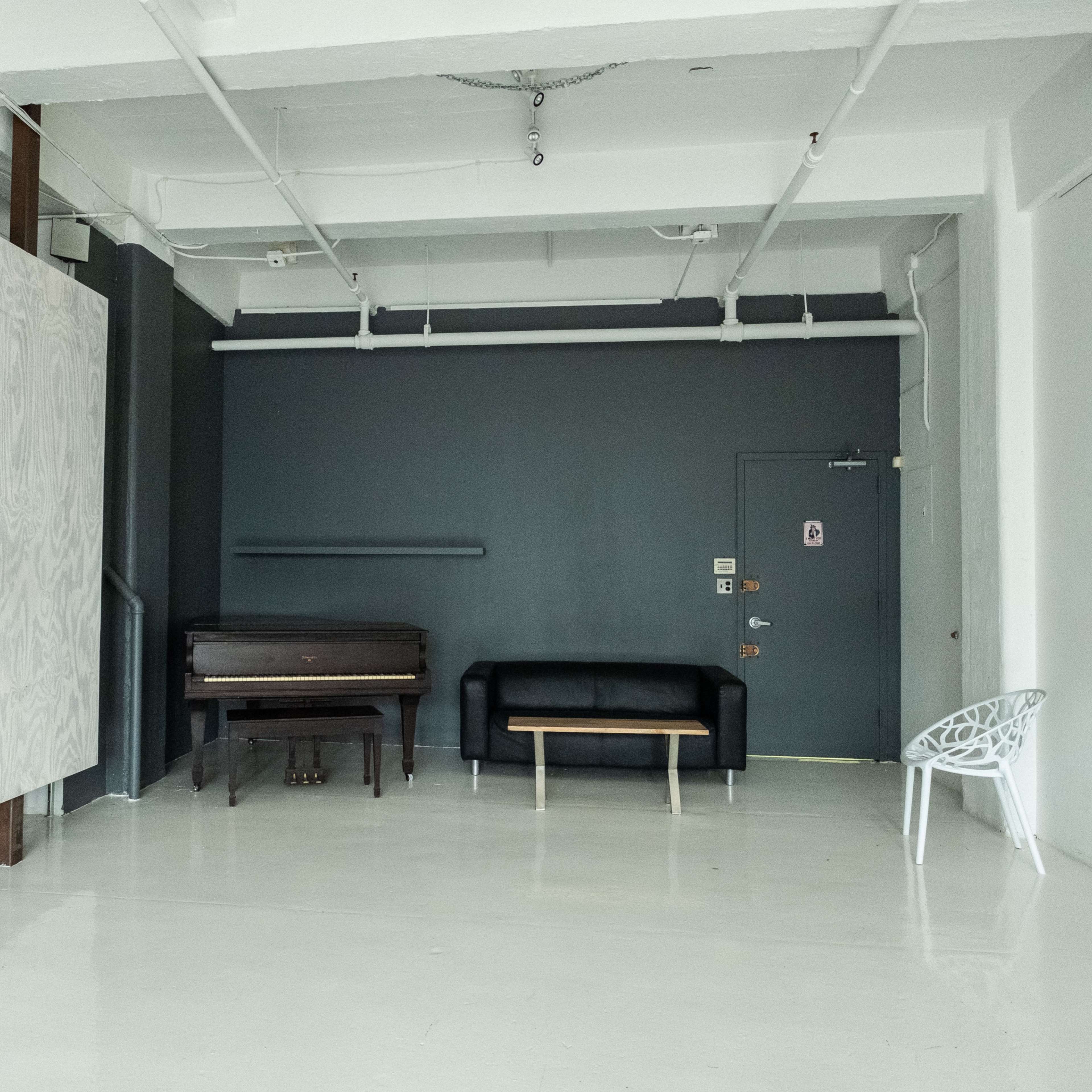A minimalist interior space featuring a black couch, a wooden bench, a piano, and a white chair against a gray wall.