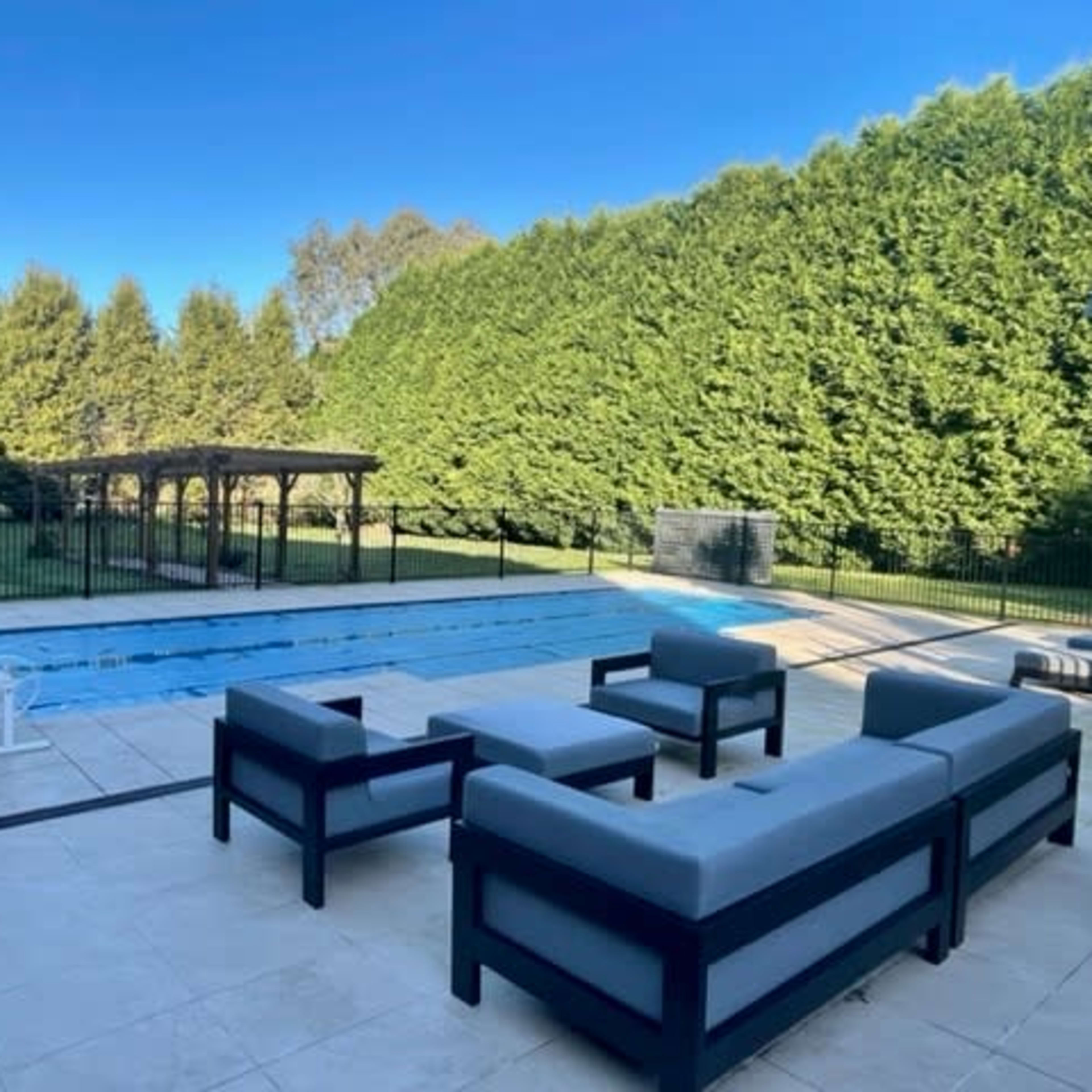 A rectangular pool with a surrounding patio and seating area is set against a backdrop of tall green hedges.