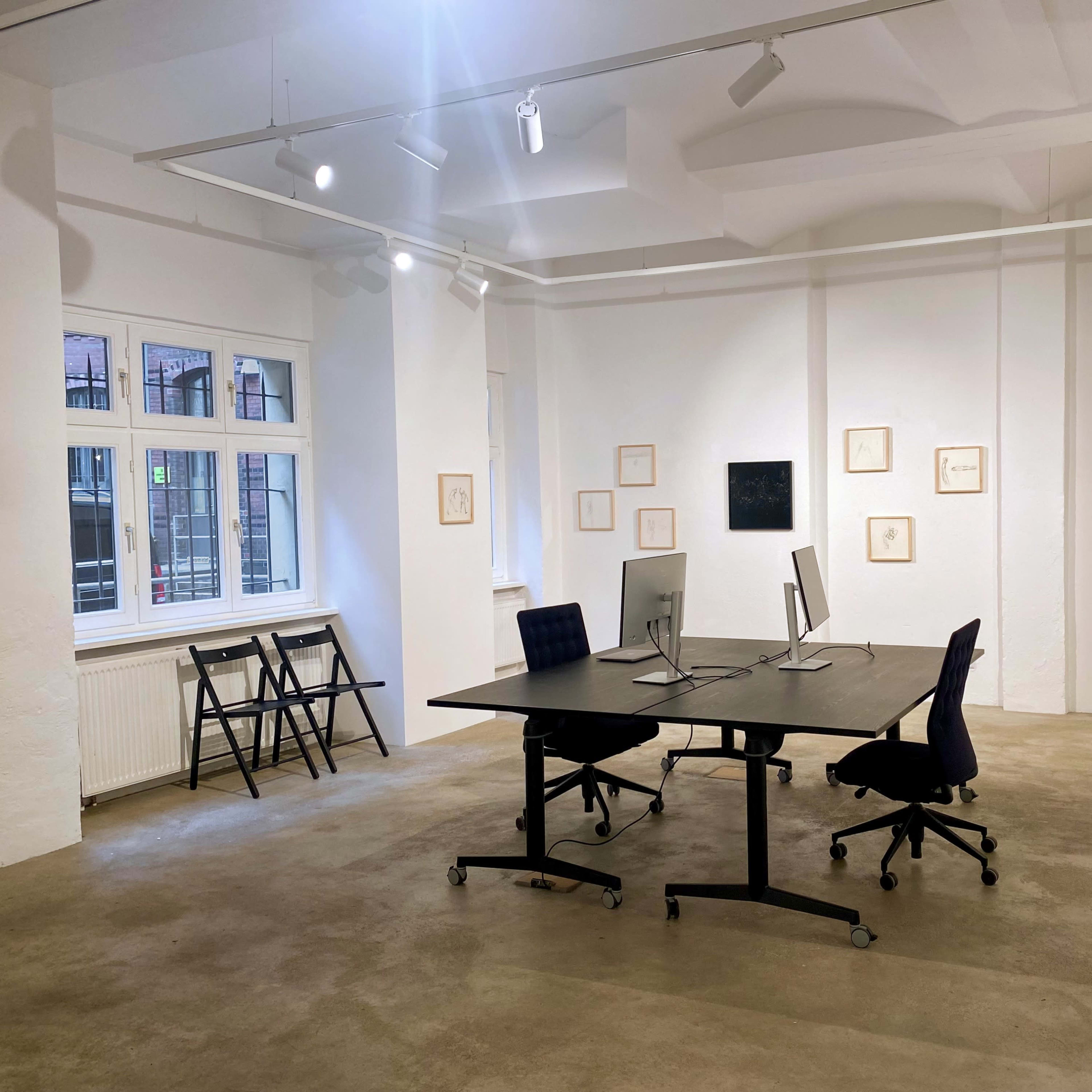 A minimalist office space featuring a large table with two monitors, surrounded by empty chairs, and artwork hanging on the white walls.