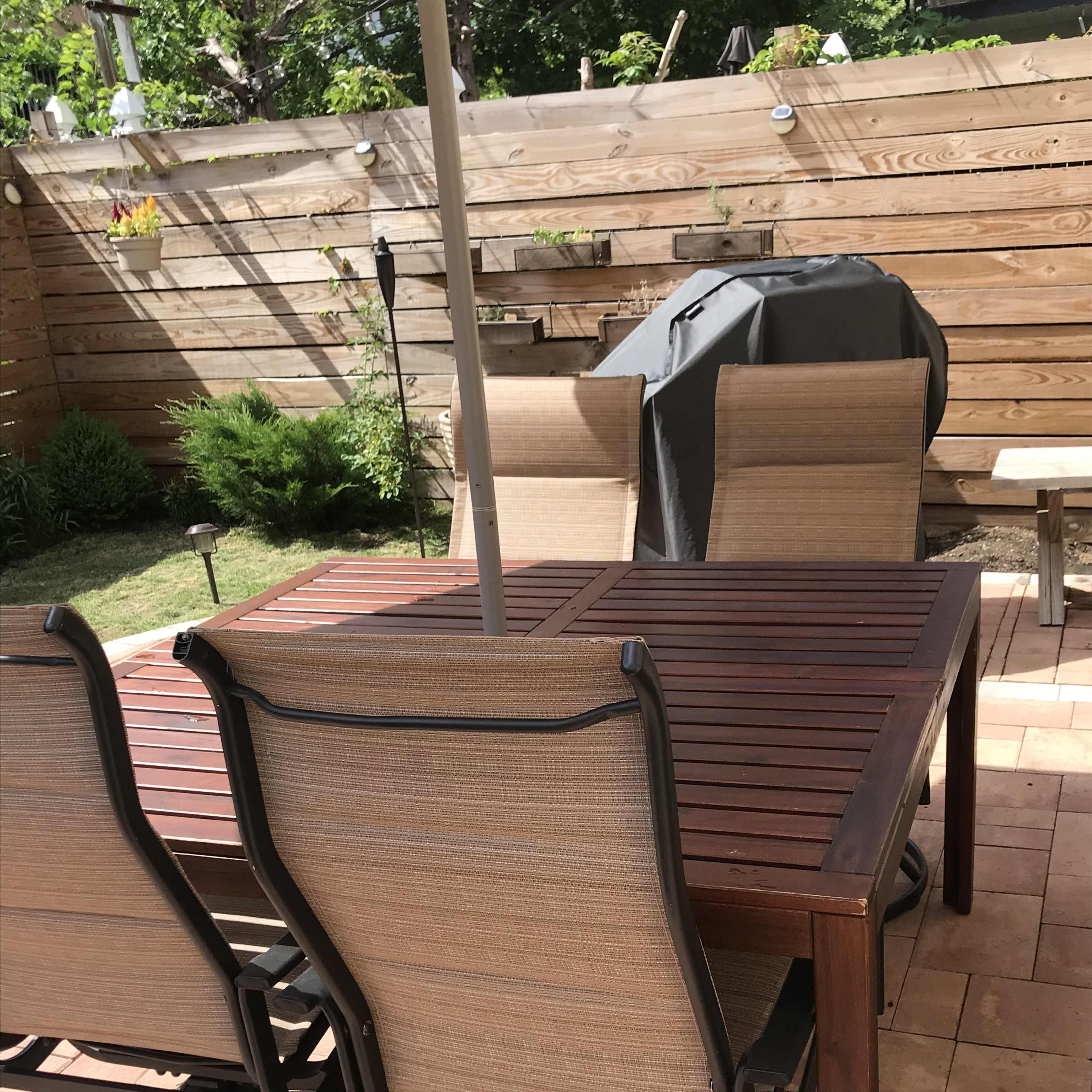 A wooden patio table with four rocking chairs sits on a tiled surface in an outdoor space surrounded by greenery and a barbecue grill.