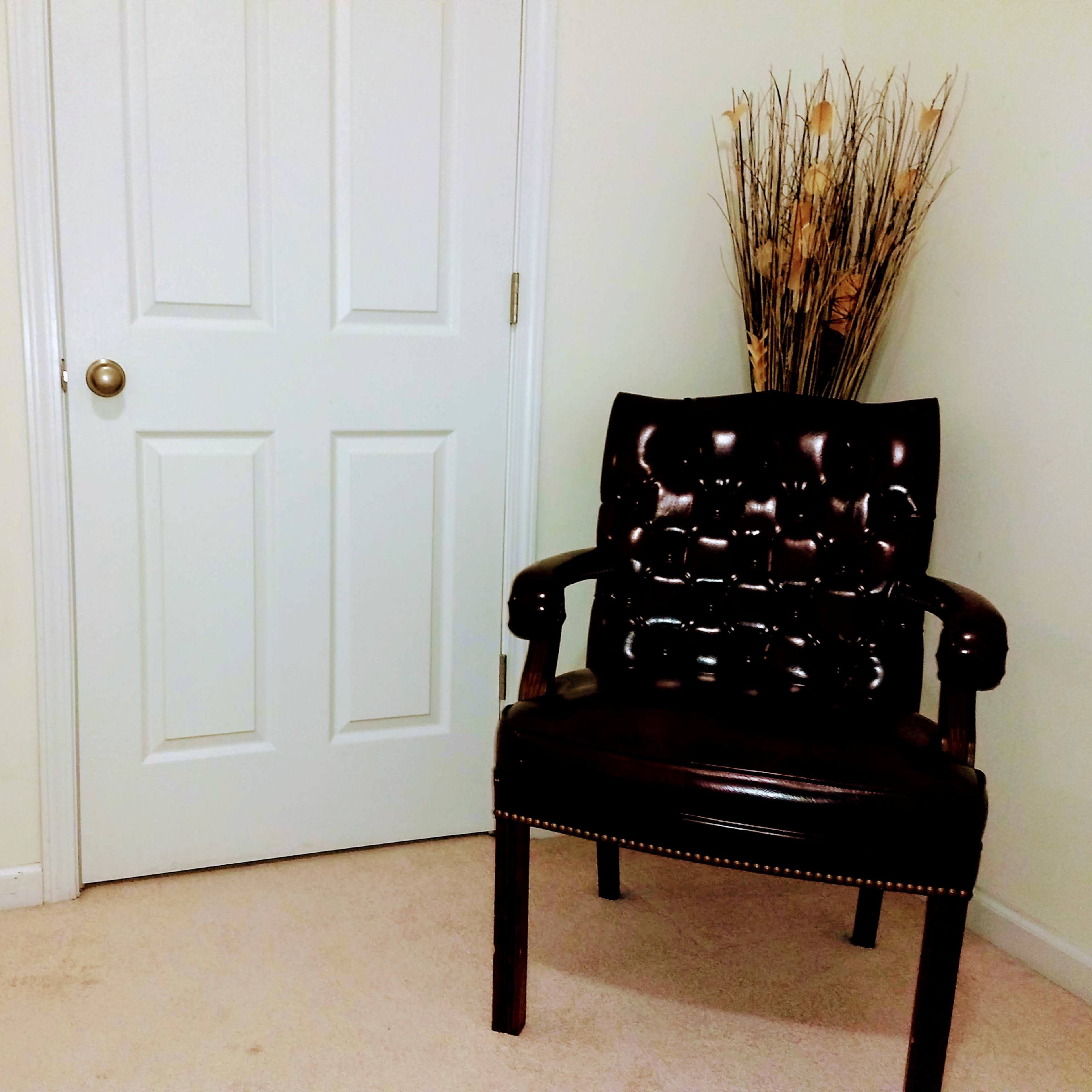 A brown upholstered chair is positioned near a closed white door, with a decorative vase of dried plants placed behind it.