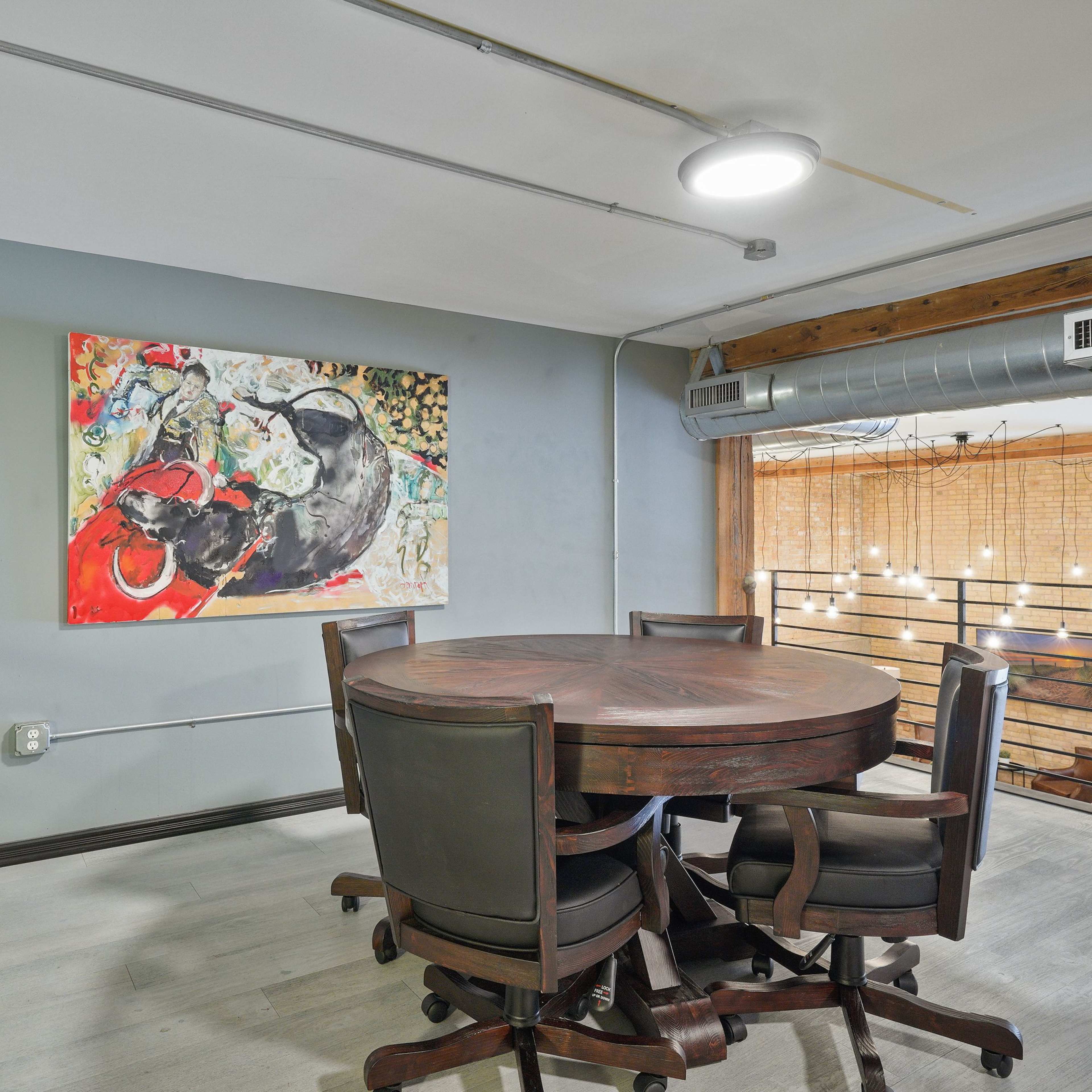 A round wooden table with dark chairs is set against a wall featuring a vibrant painting, in an interior space with industrial elements and soft lighting.