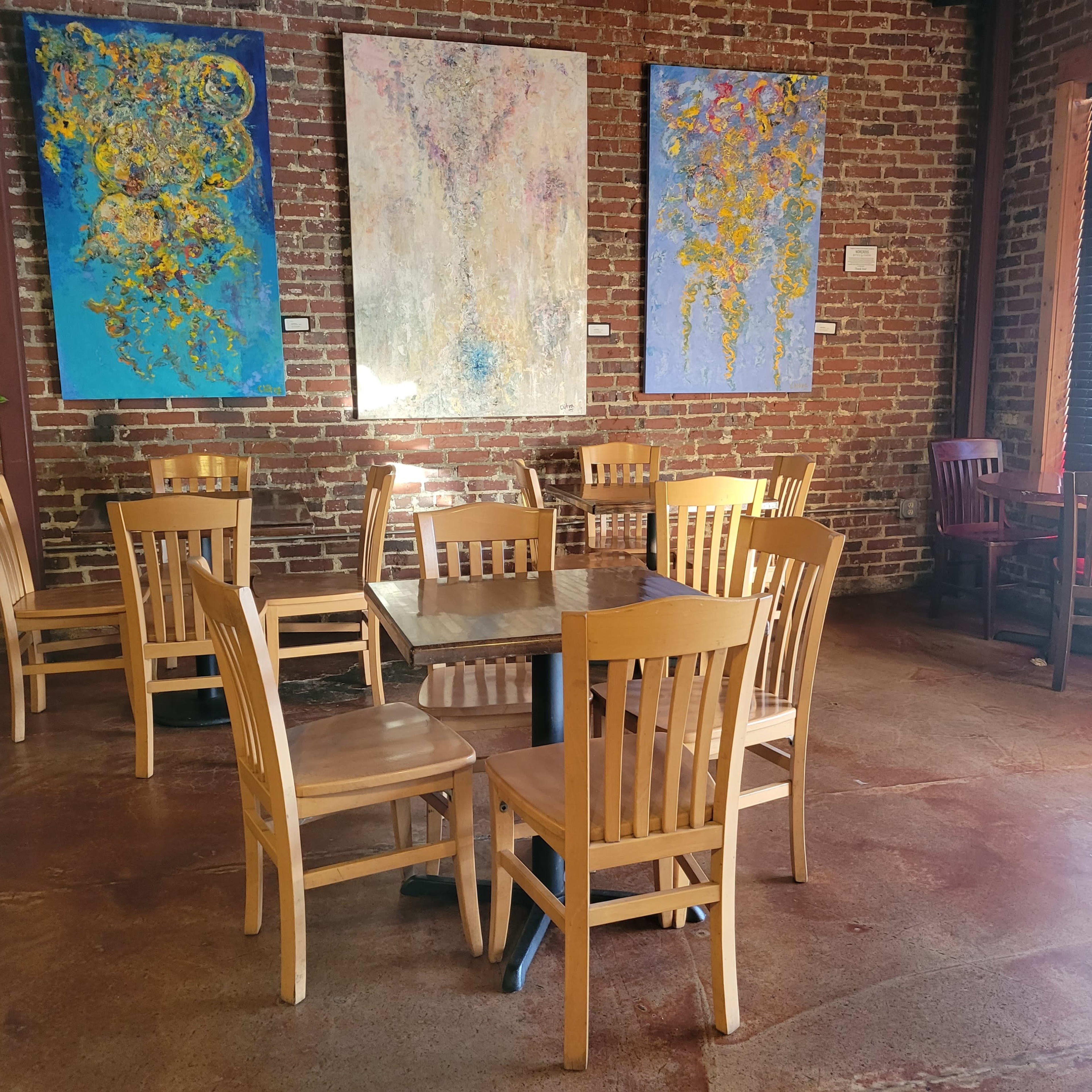 A rustic café interior with wooden tables and chairs arranged around a central table, featuring brick walls and colorful abstract paintings.
