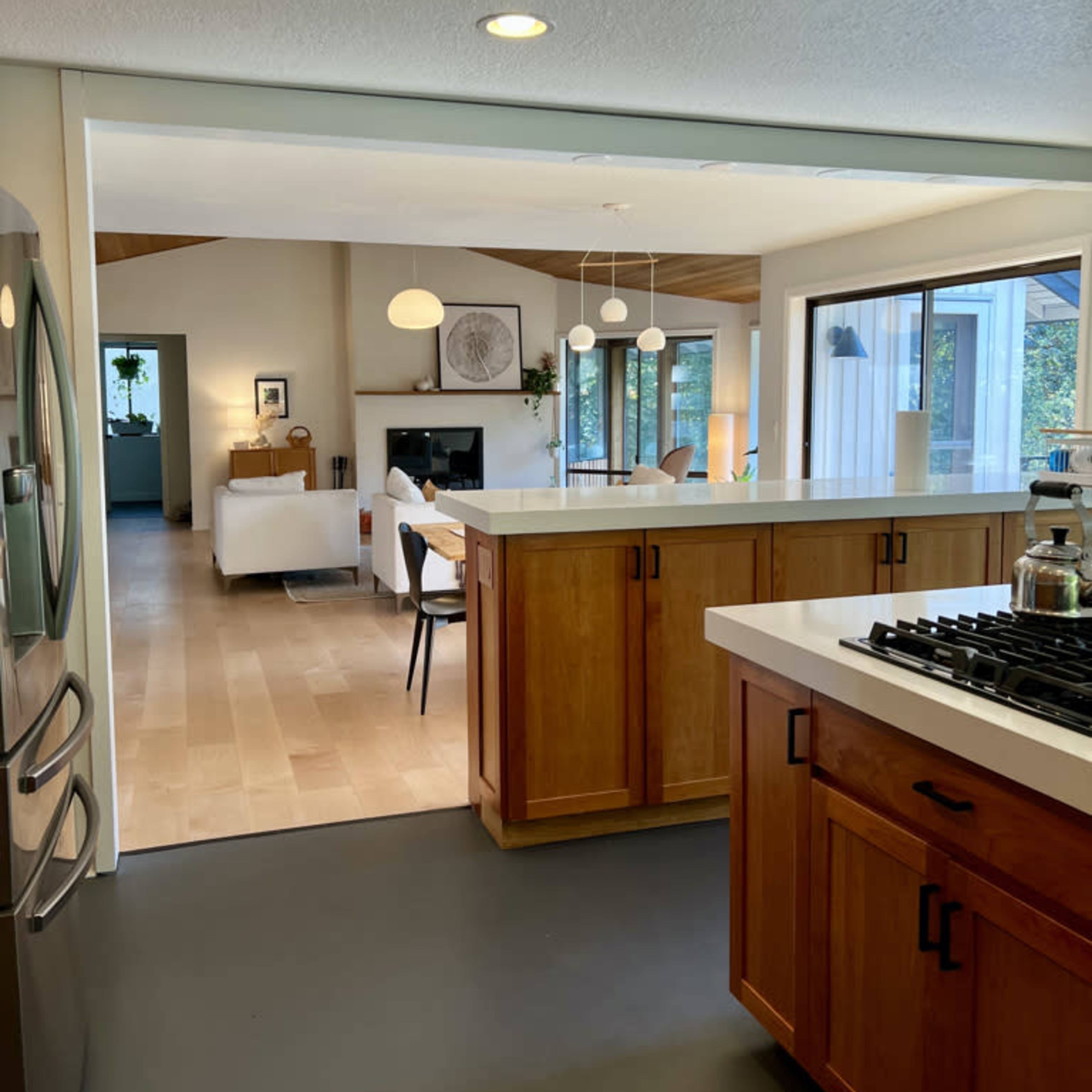 A modern kitchen with wooden cabinets and a gas stove, opening up to a bright living space with a sofa and large windows.