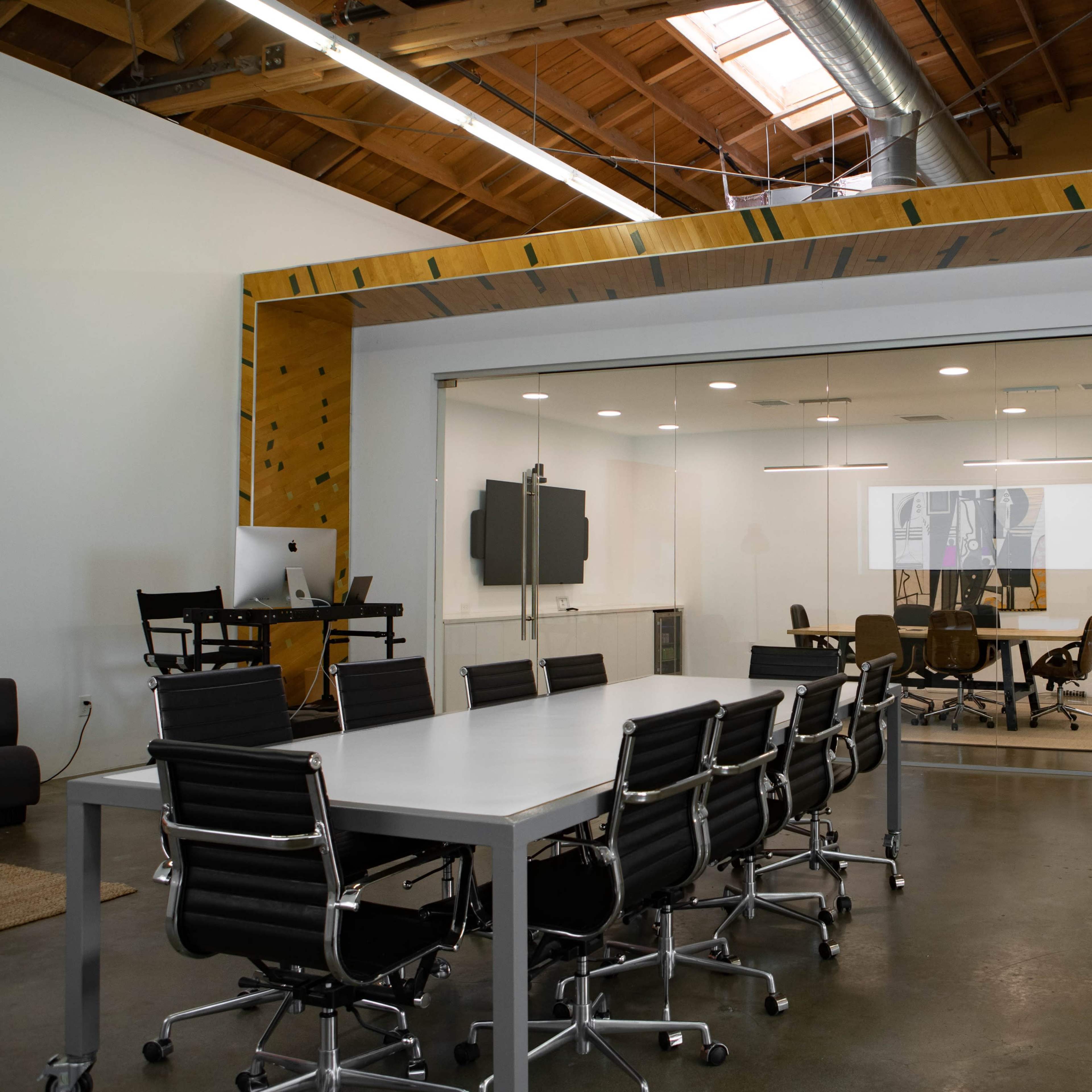A modern office space featuring a large conference table surrounded by black chairs, with a glass-walled meeting room visible in the background.
