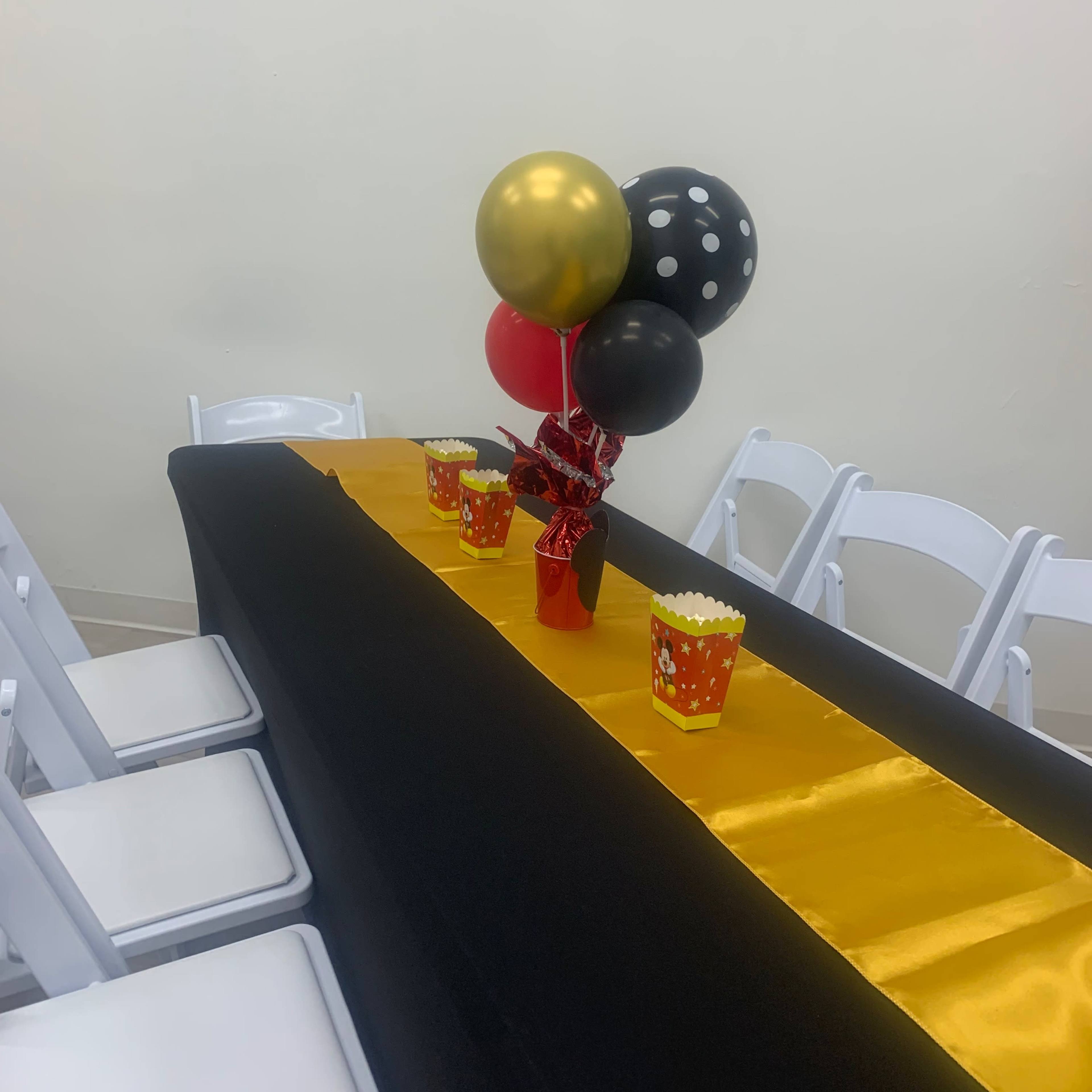 A table is set with a black tablecloth, a gold runner, popcorn containers, and balloons in black, gold, and red.