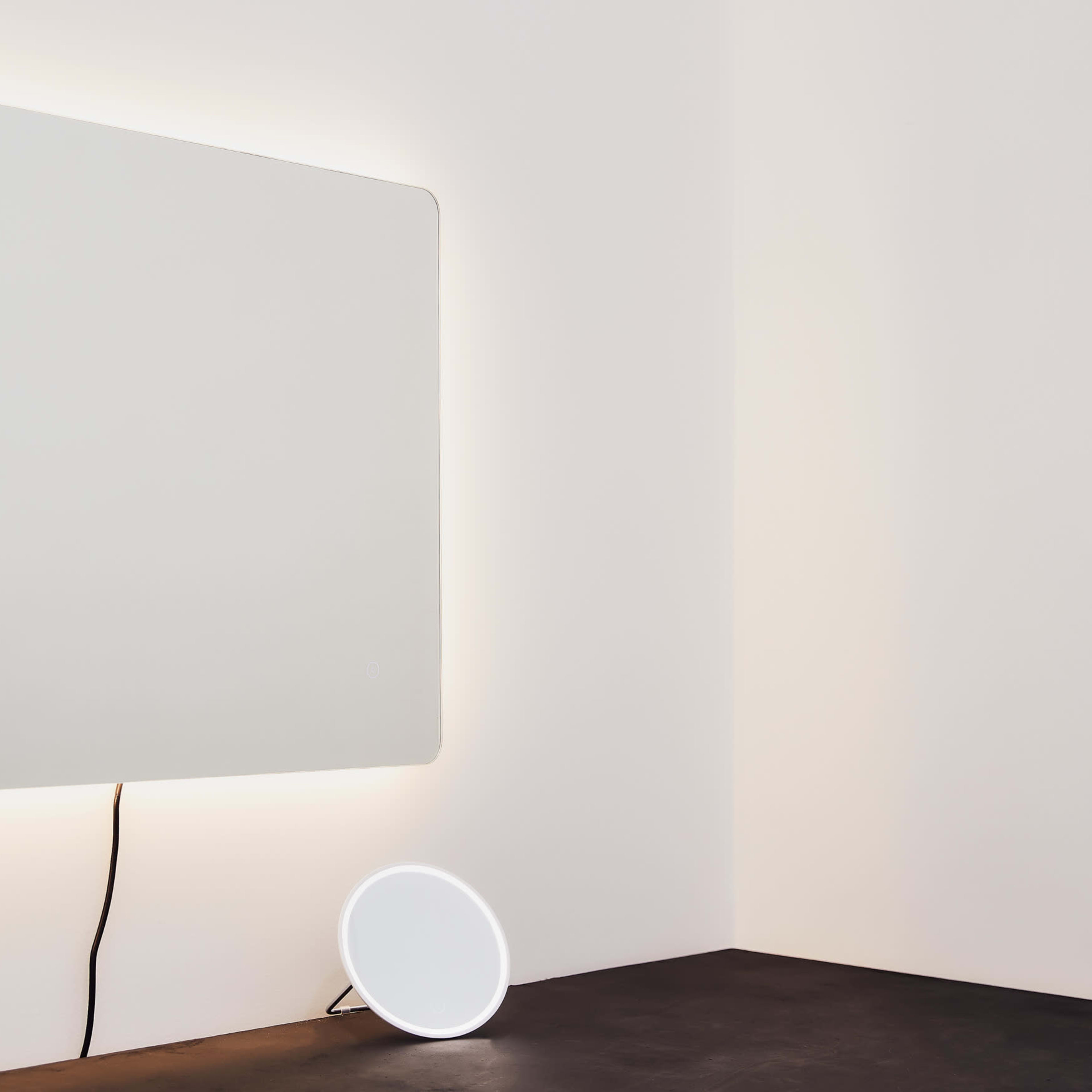 A wall-mounted mirror with backlighting is positioned above a small round mirror on a black surface in a softly lit room.