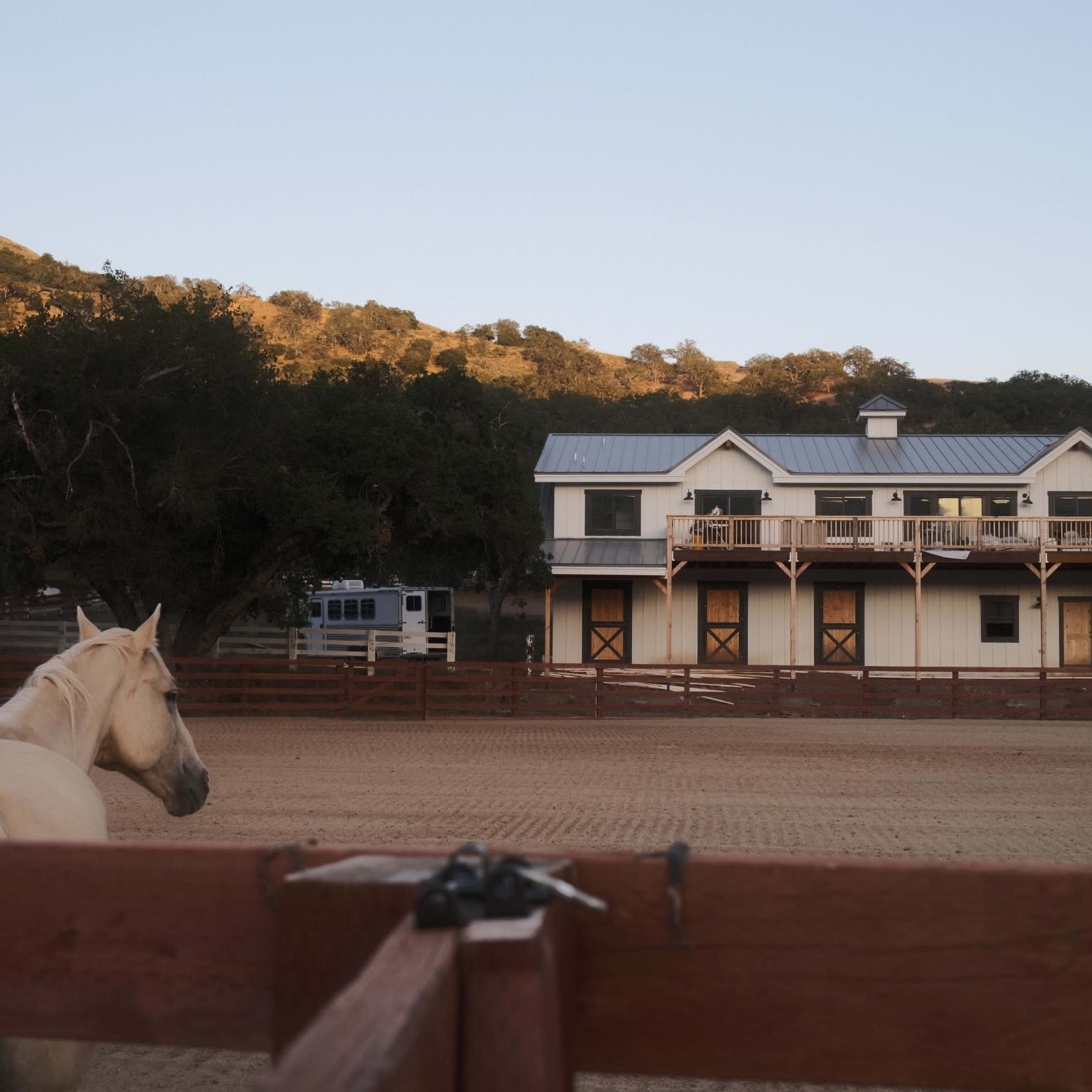 A horse stands near a wooden fence while a two-story stable is visible in the background against a hillside.