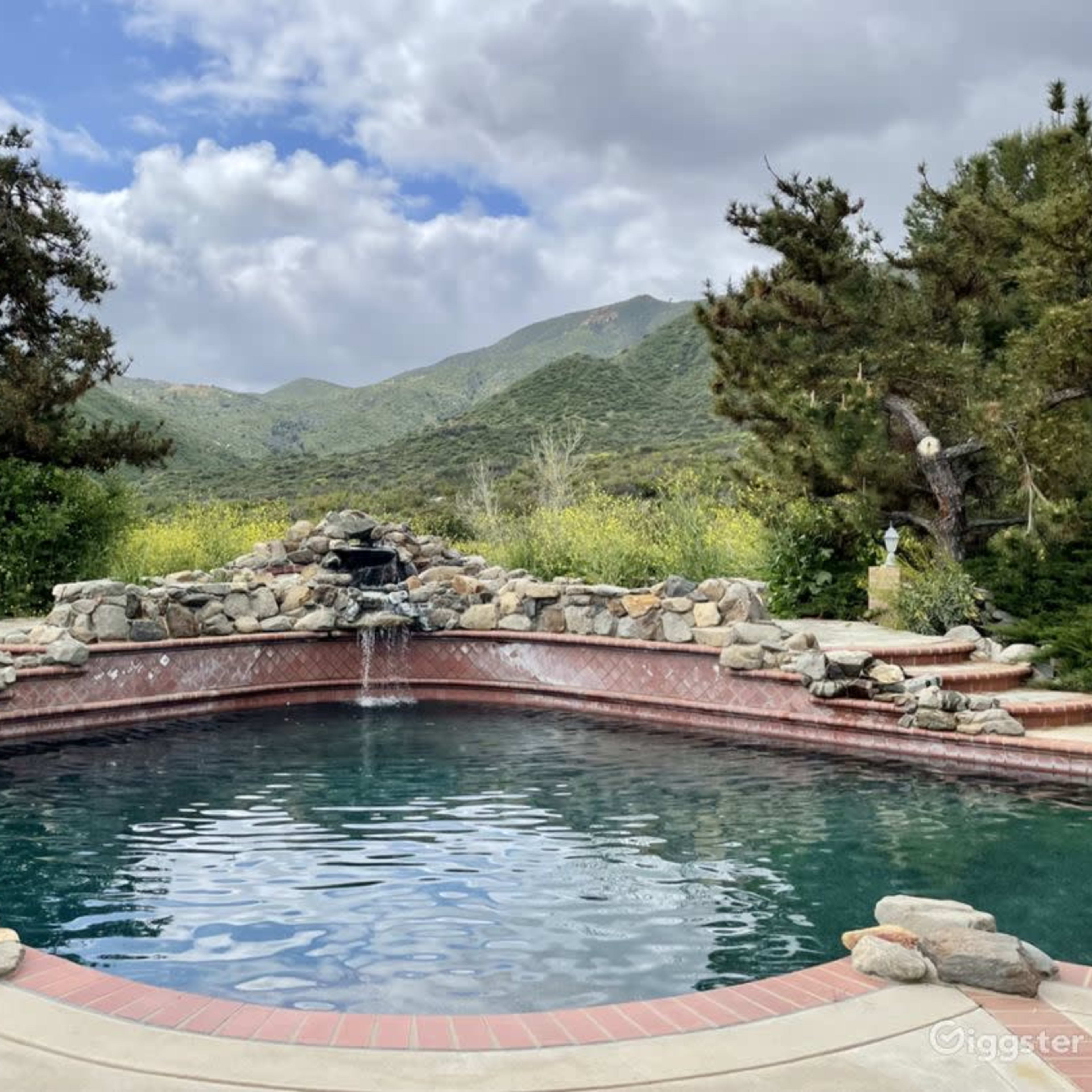 A stone-lined swimming pool with a waterfall and mountain scenery in the background under a partly cloudy sky.