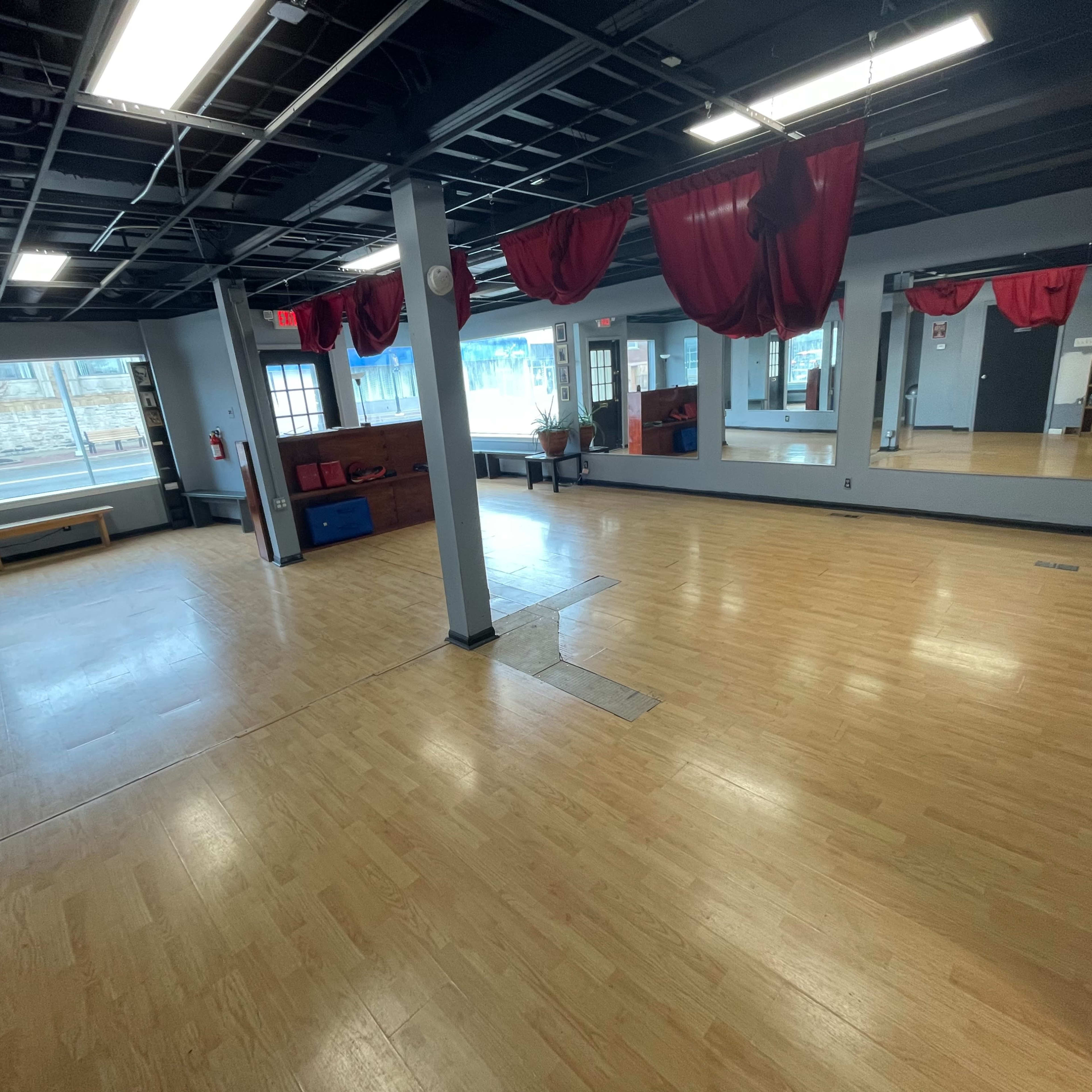 A spacious, empty studio with wooden floors, large mirrors, and red drapes hanging from the ceiling.