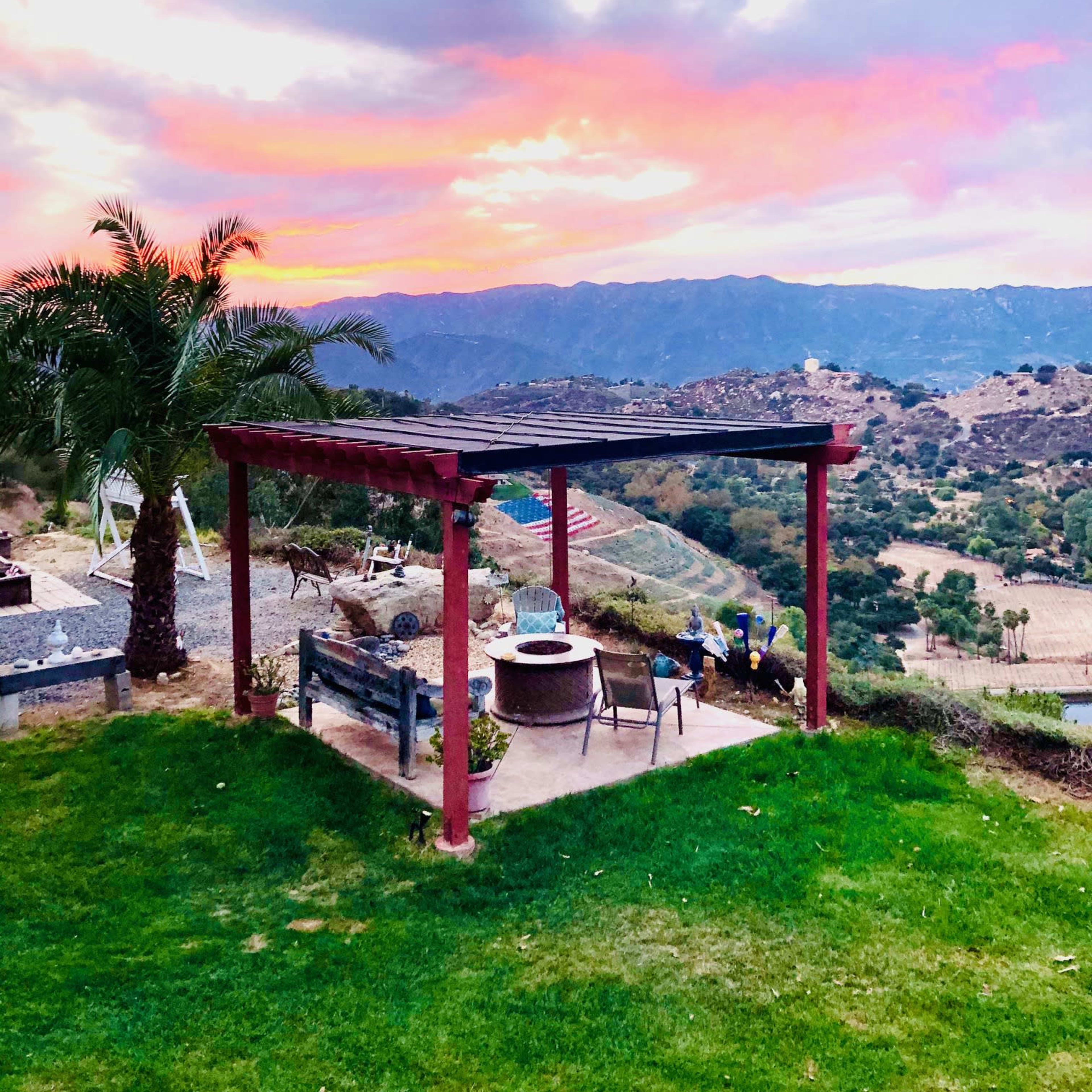 A shaded seating area with a table and chairs is set against a backdrop of mountains and a colorful sunset sky.