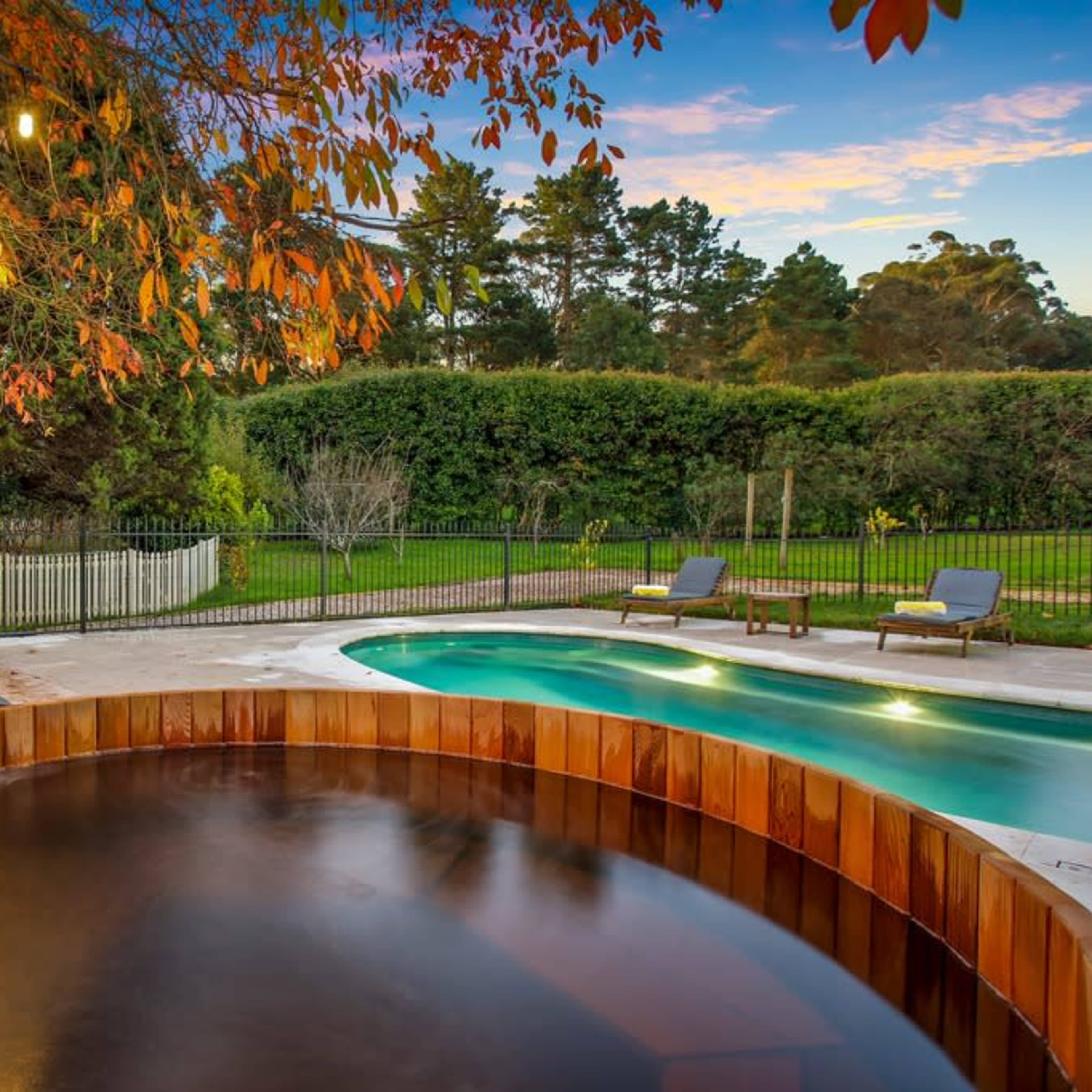 A wooden hot tub next to a swimming pool, surrounded by lush greenery and softly lit by hanging lights.