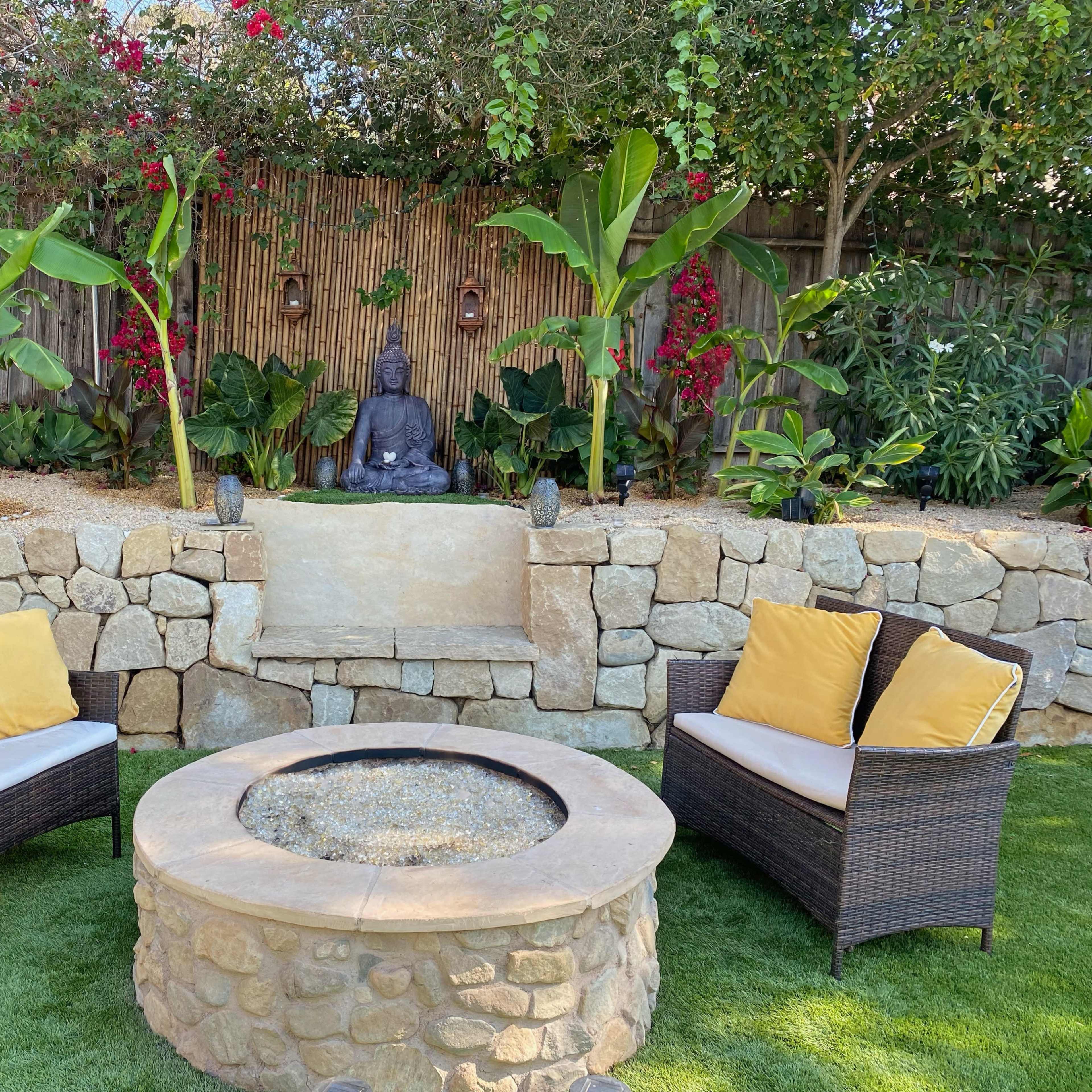 A cozy outdoor seating area features two wicker chairs with yellow cushions, facing a stone fire pit and a decorative Buddha statue surrounded by lush plants and vibrant flowers.