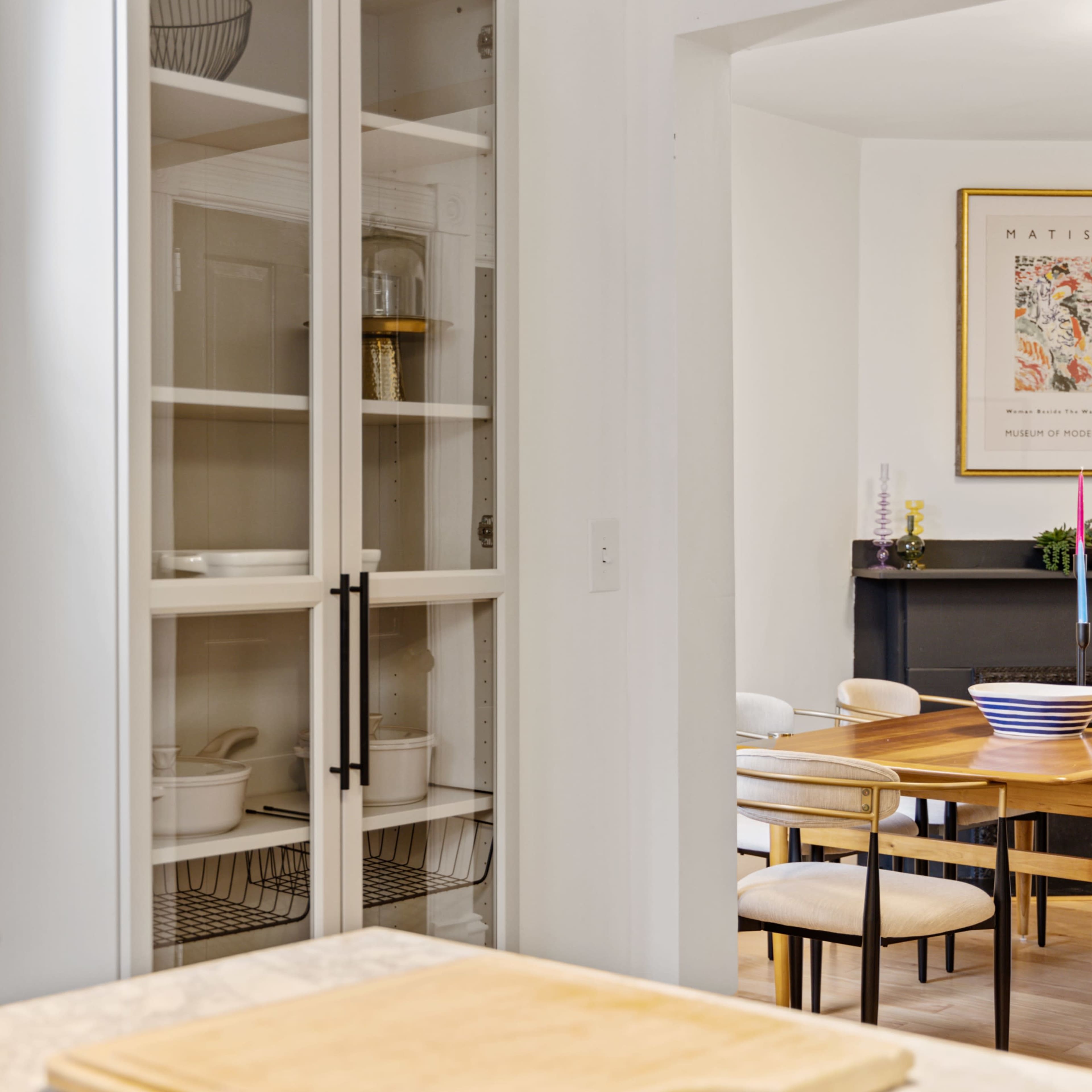 A modern kitchen featuring a clear glass-front pantry cabinet and a dining area in the background with wooden tables and chairs.