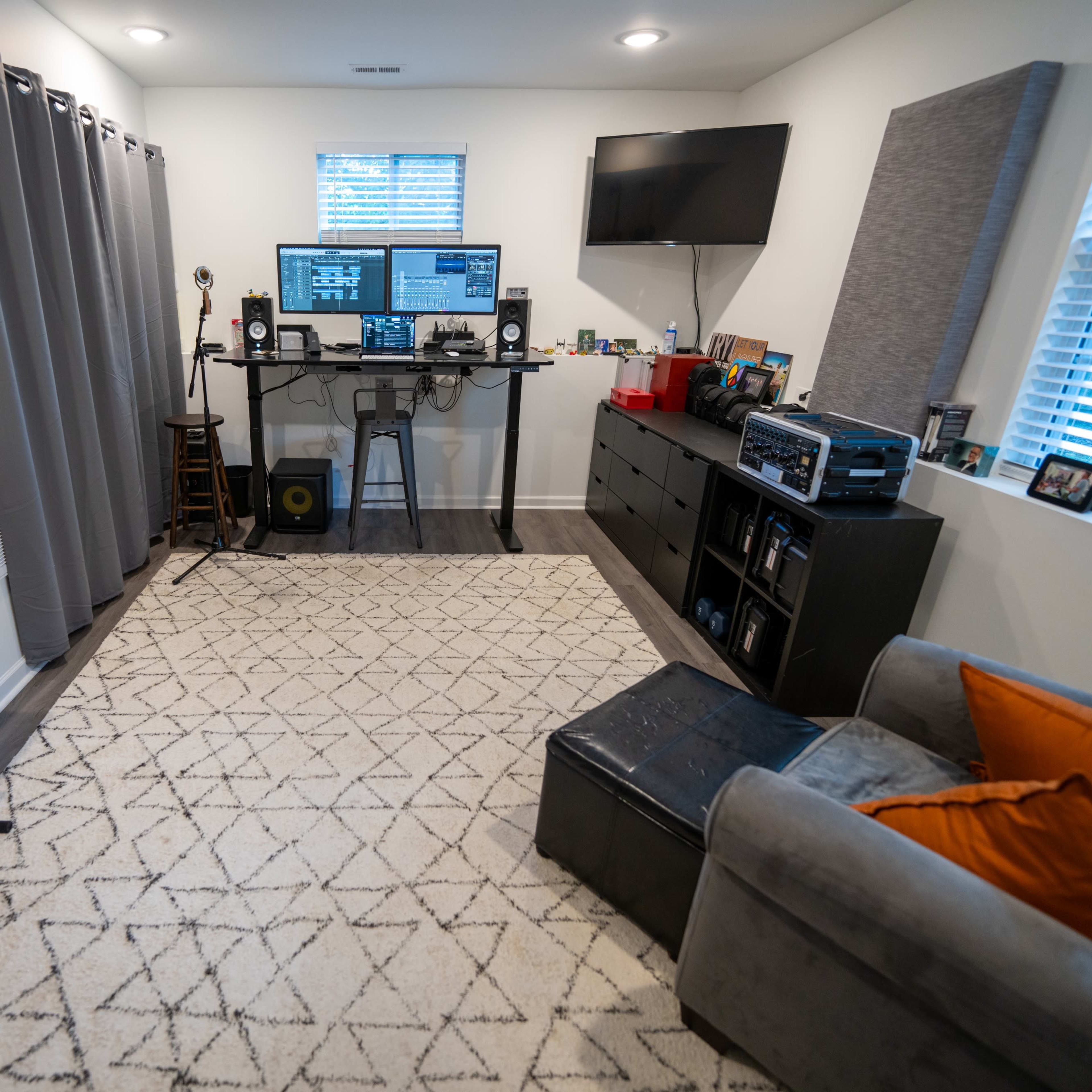 A home studio setup with a computer desk, sound equipment, a keyboard, and a gray couch on a patterned rug.
