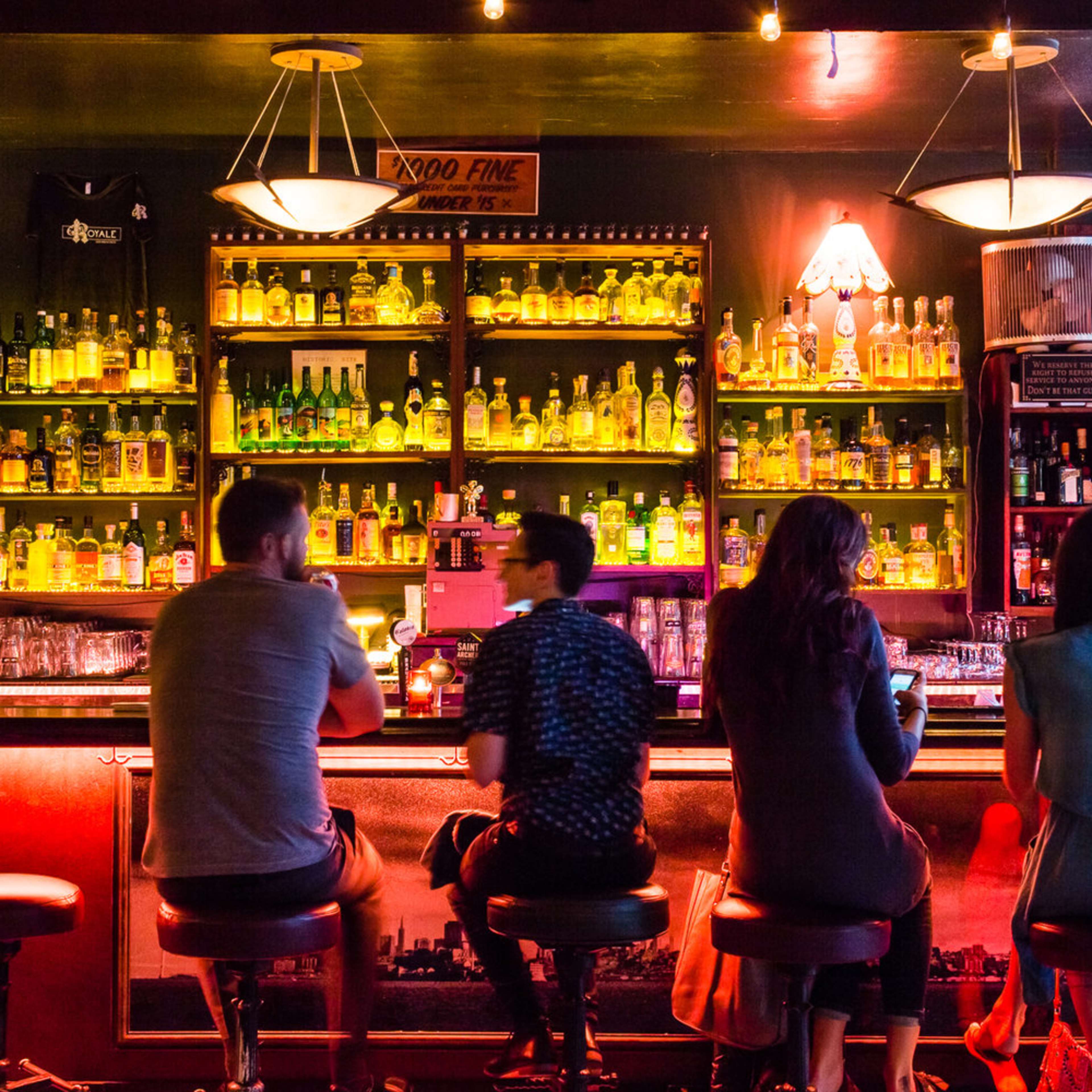 A group of people sits at a bar, facing a illuminated shelf filled with various bottles.
