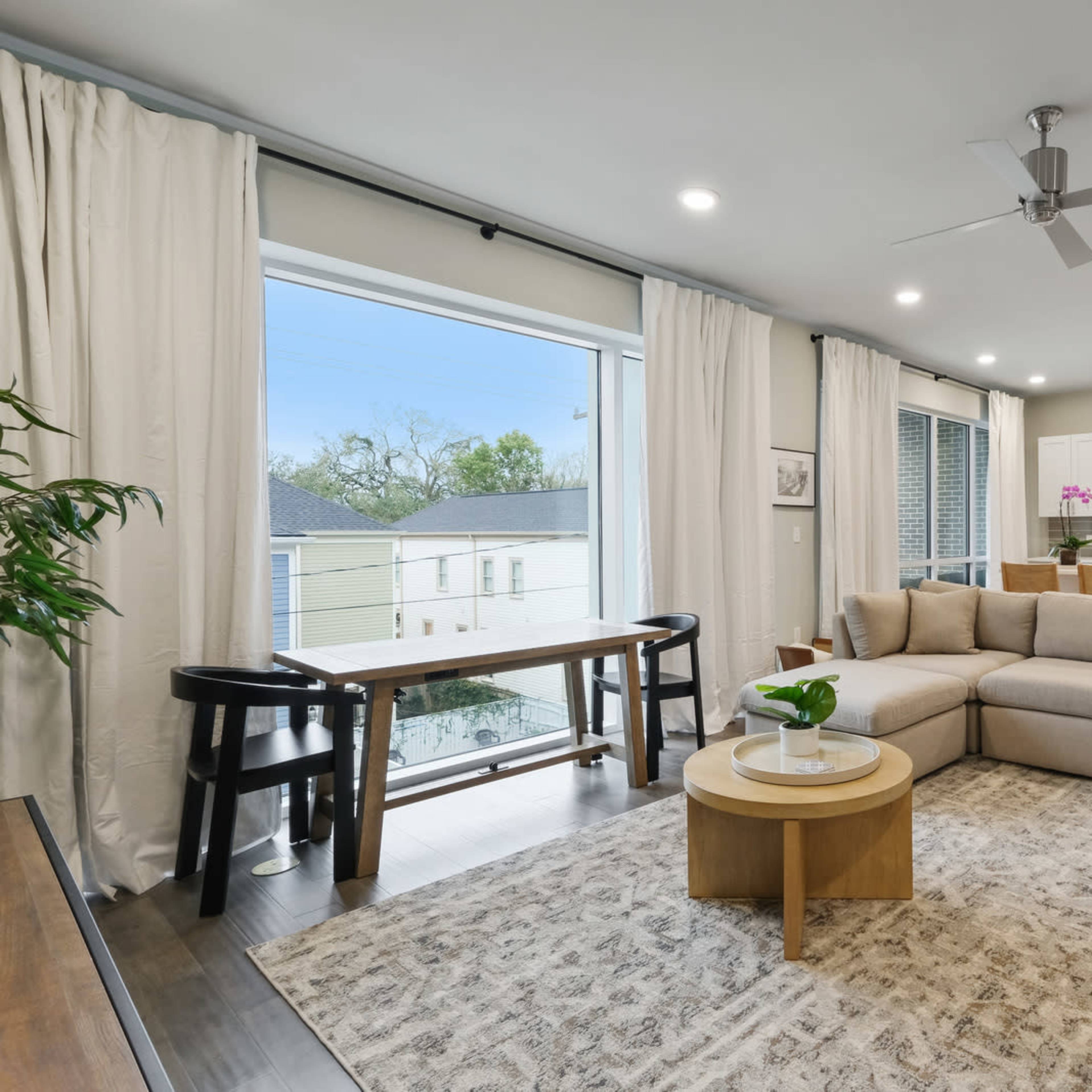 A modern living room features a large sectional sofa, a round coffee table, and a spacious kitchen in the background, with large windows allowing natural light to enter the space.