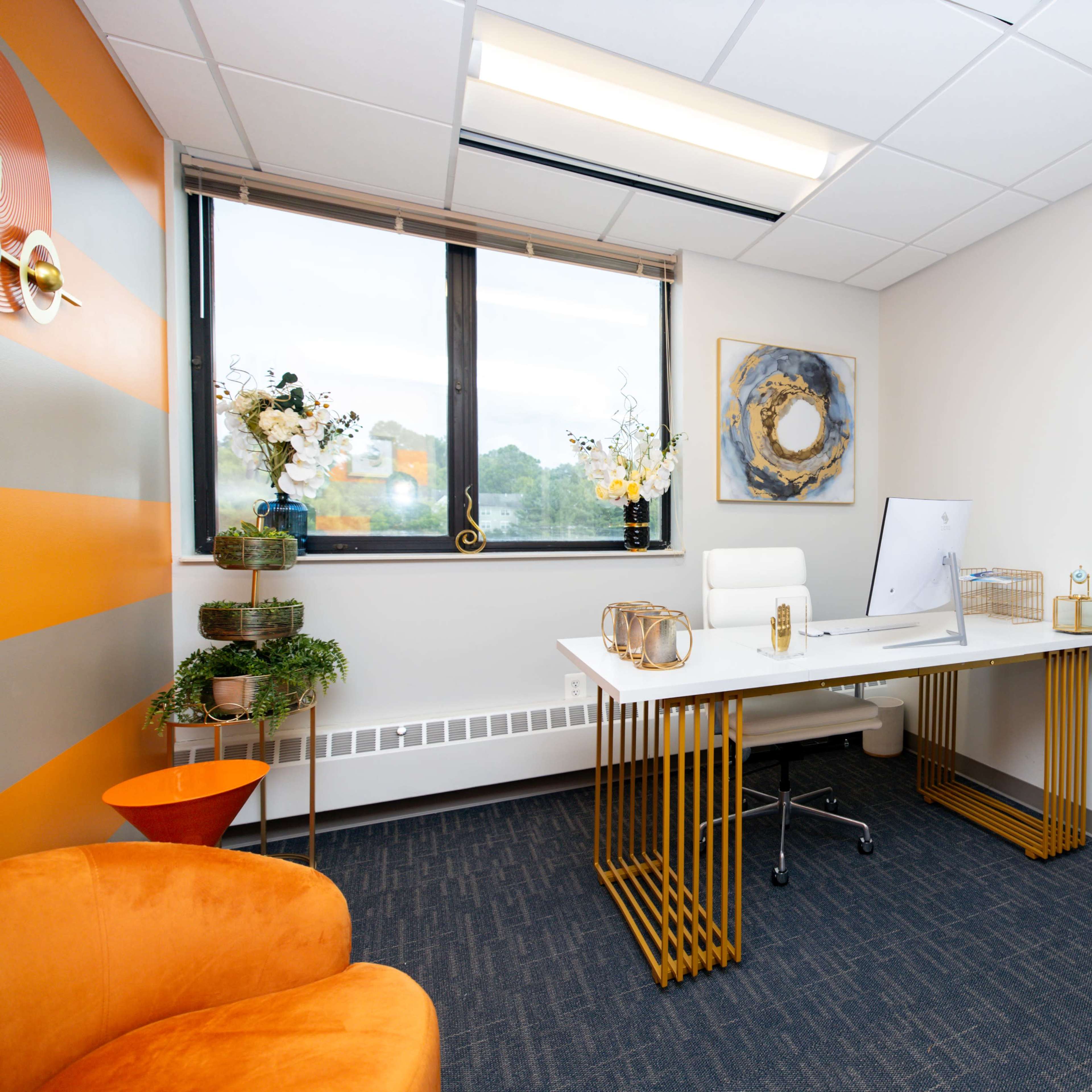 A modern office space featuring a white desk with a computer, an orange chair, and decorative plants on the windowsill.