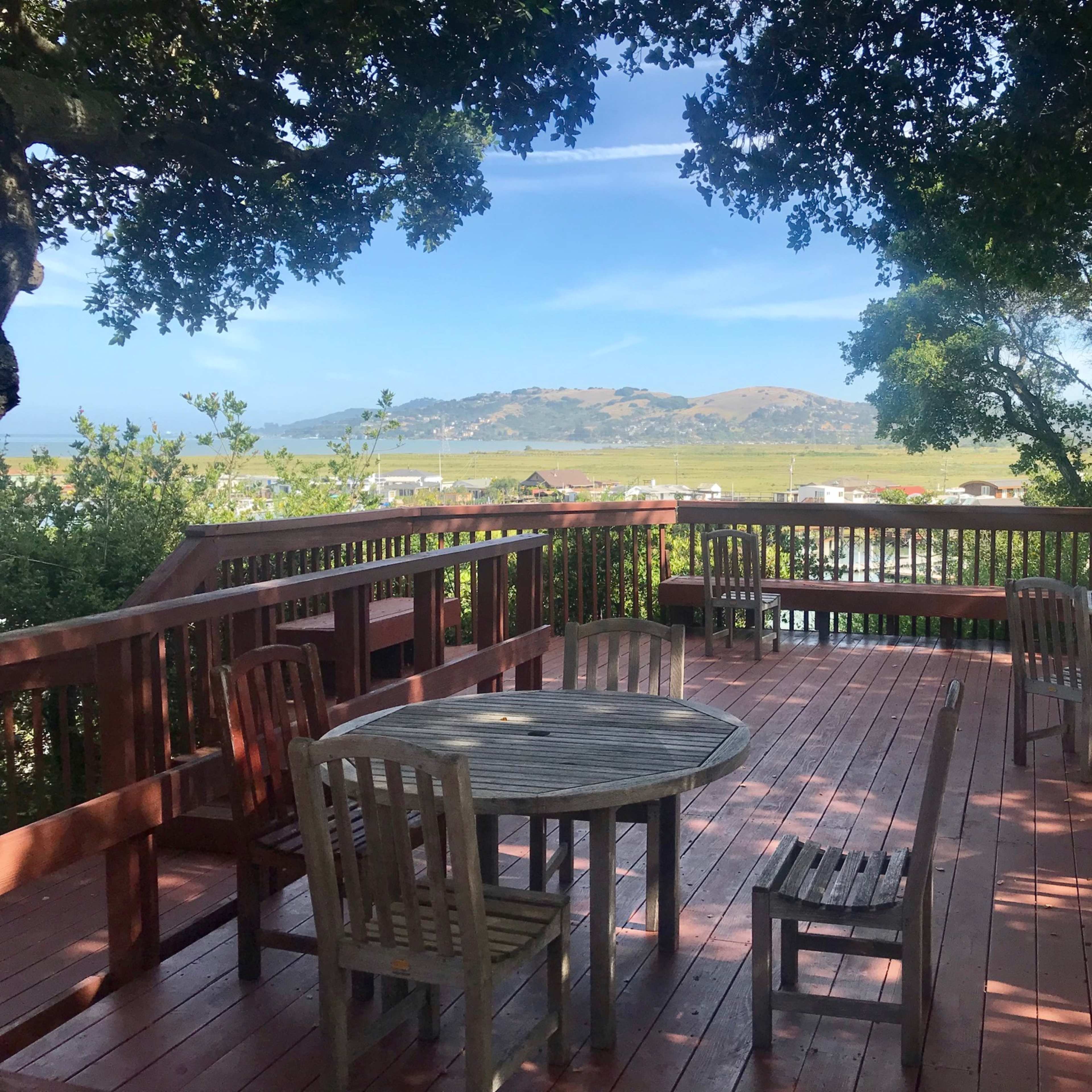 A wooden deck with several tables and chairs overlooks a landscape of hills and trees under a clear sky.