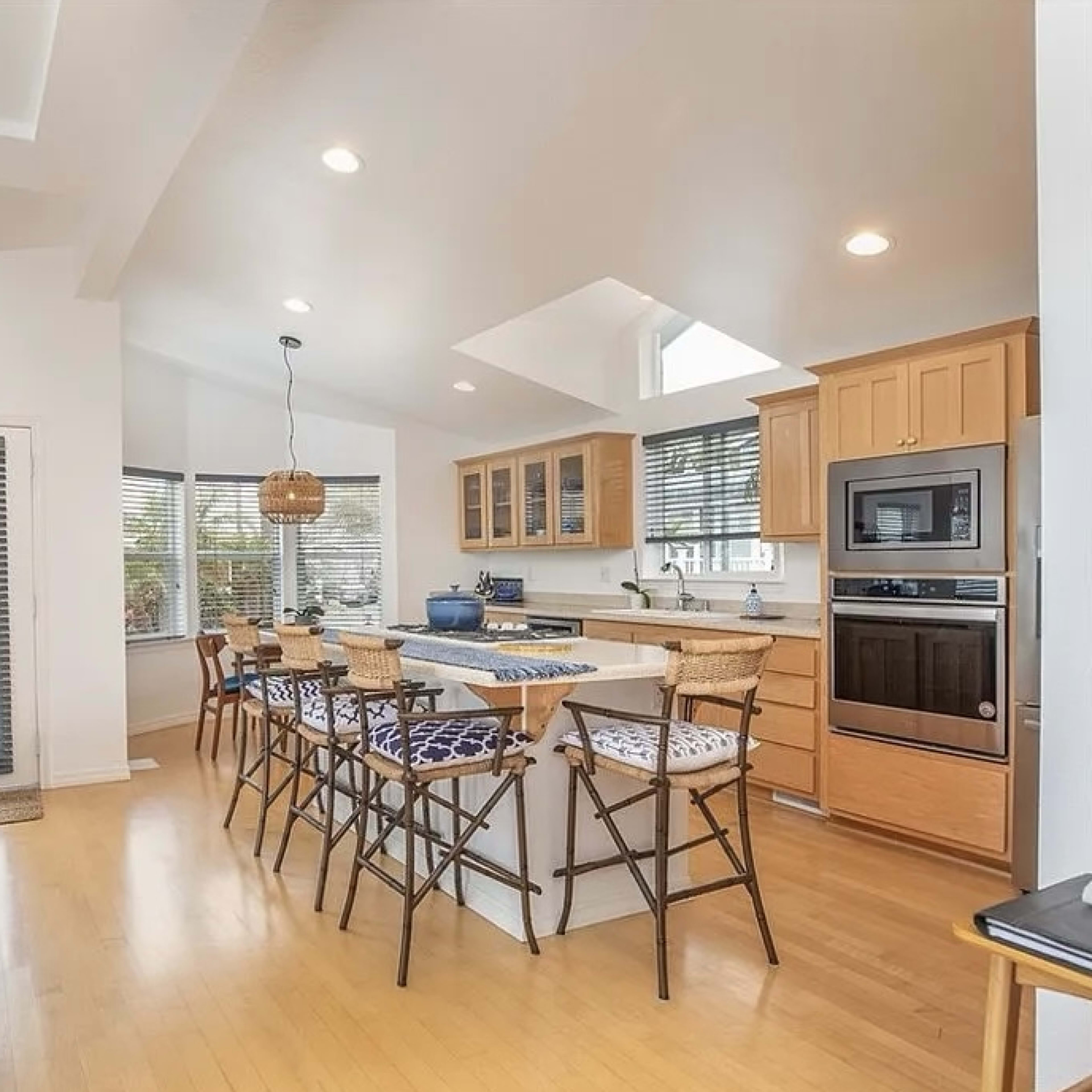 A well-lit kitchen and living area feature a central island with seating, wooden cabinetry, and large windows.