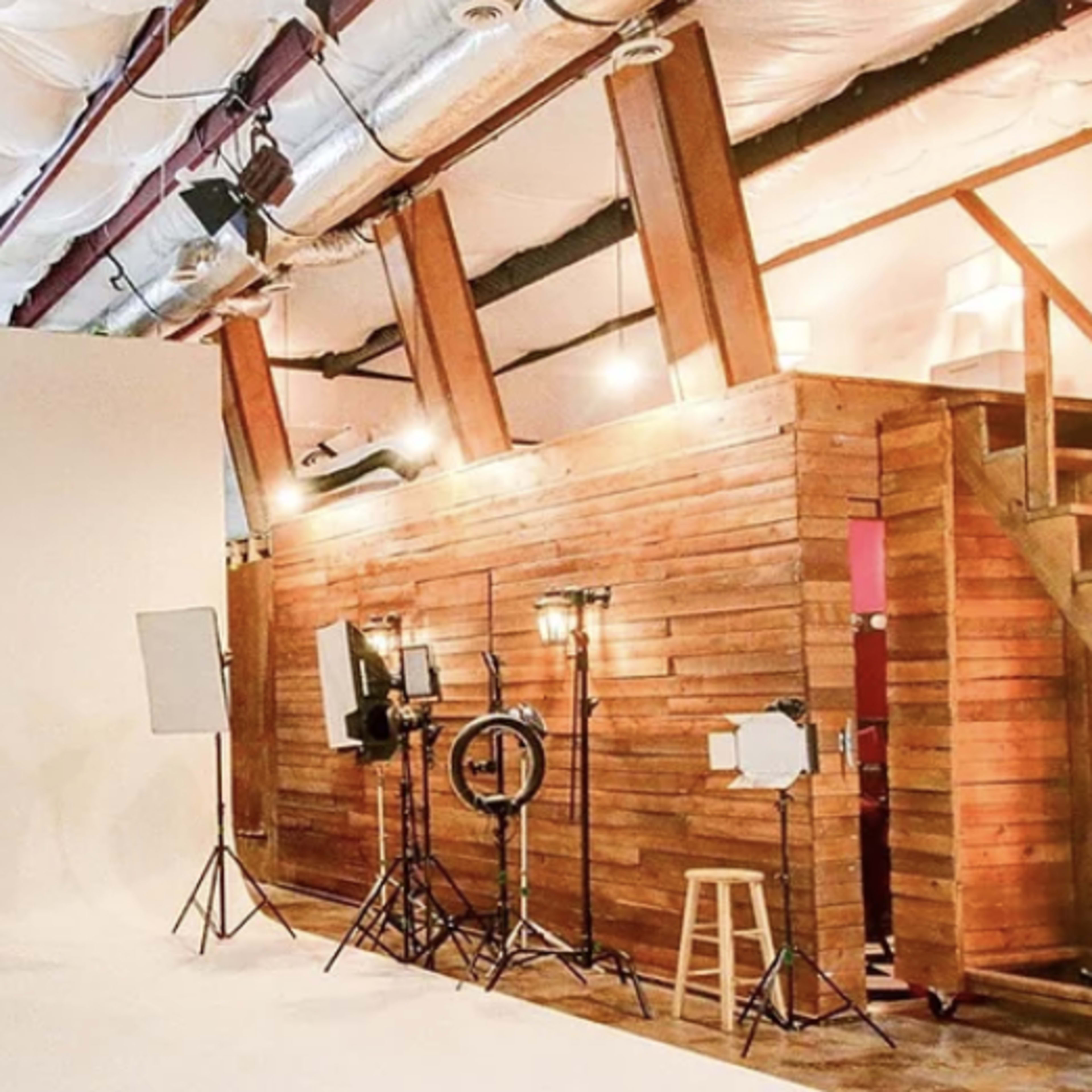 A photography studio with a white backdrop, wooden walls, lighting equipment, and a staircase leading to an upper level.