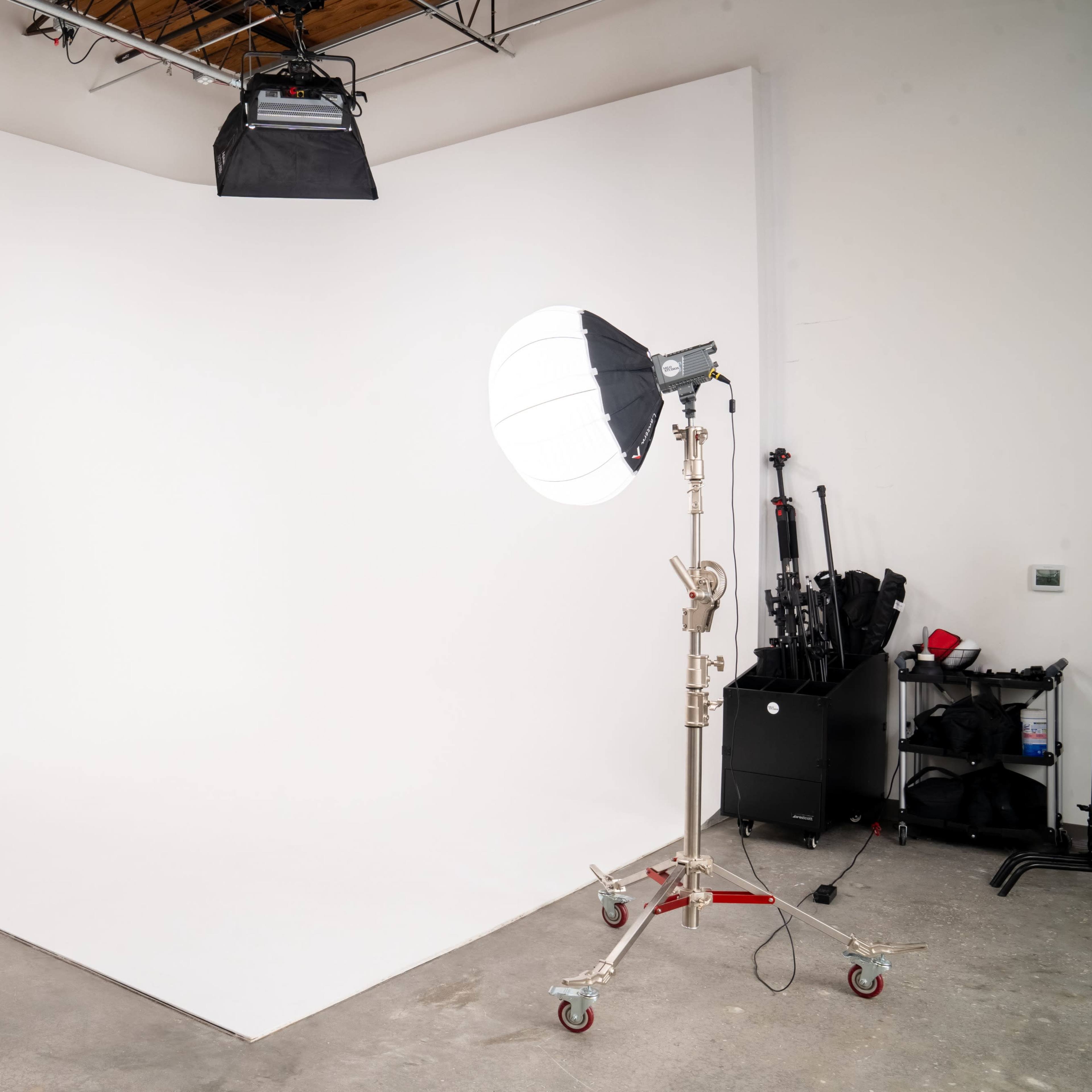 A photography studio with a white backdrop, a light stand with a softbox, and various equipment arranged in the corner.