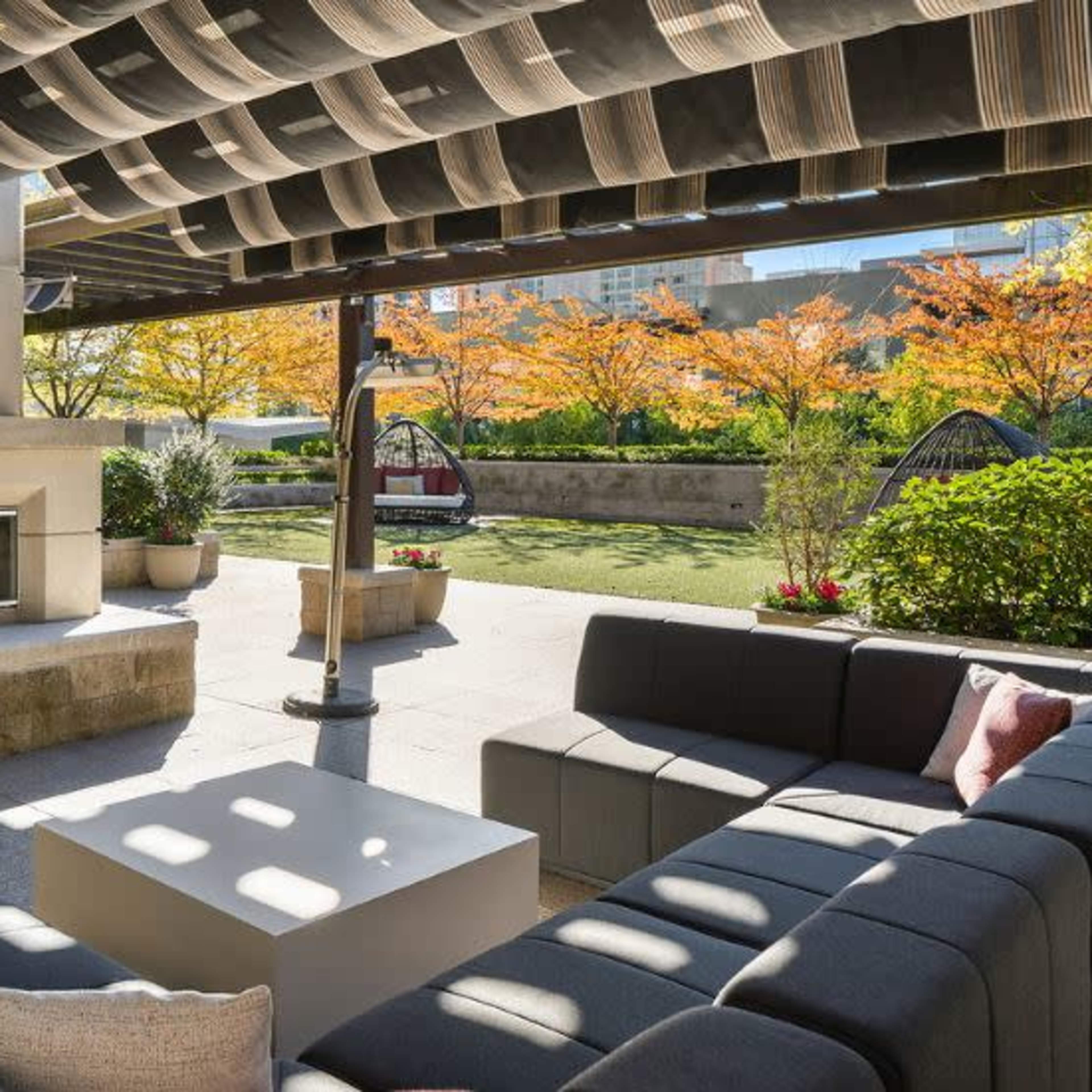 A modern outdoor lounge area with a fireplace, surrounded by greenery and benches, under a shaded pergola.