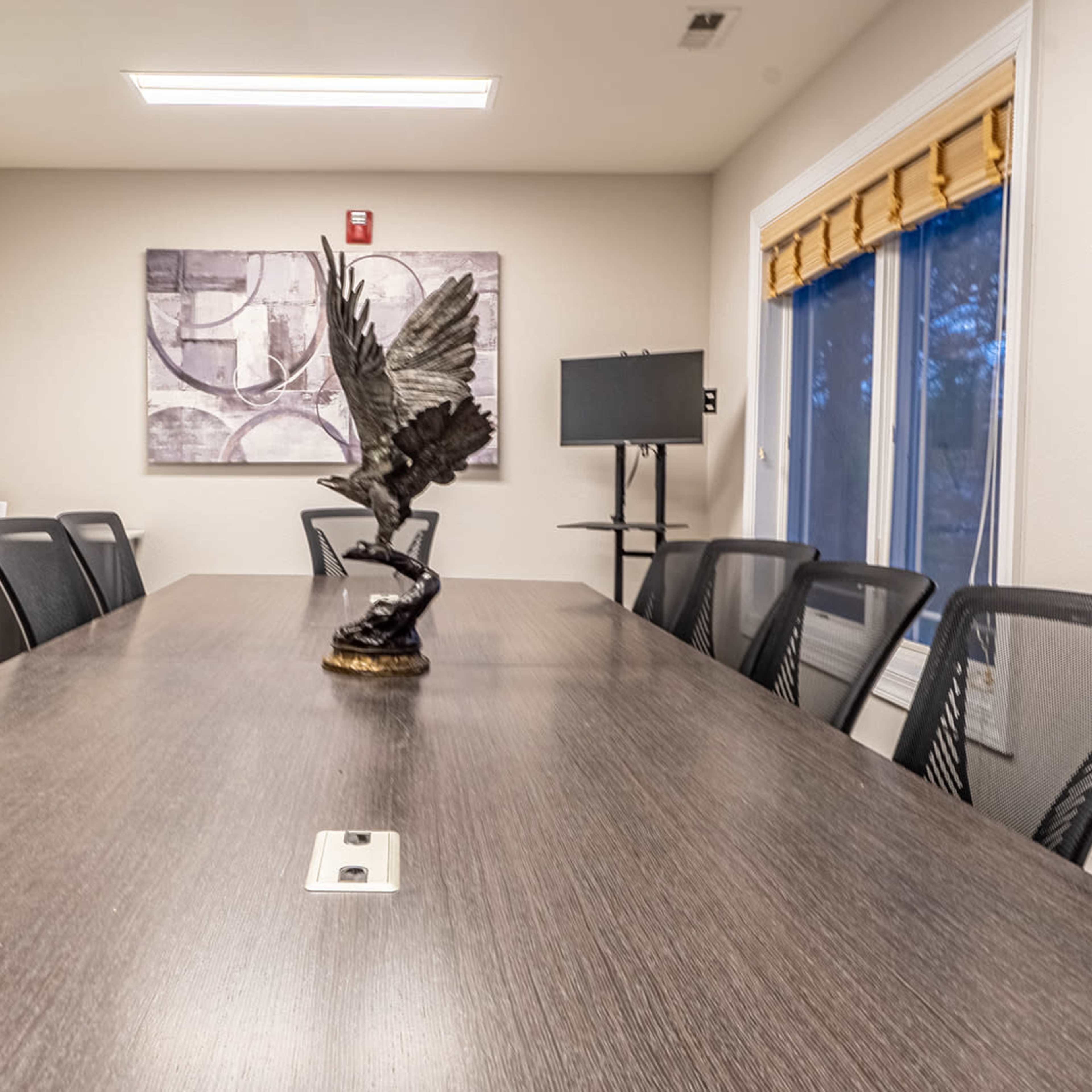 A long conference table with black chairs is set in a well-lit meeting room, featuring a decorative sculpture at its center and large windows along one wall.