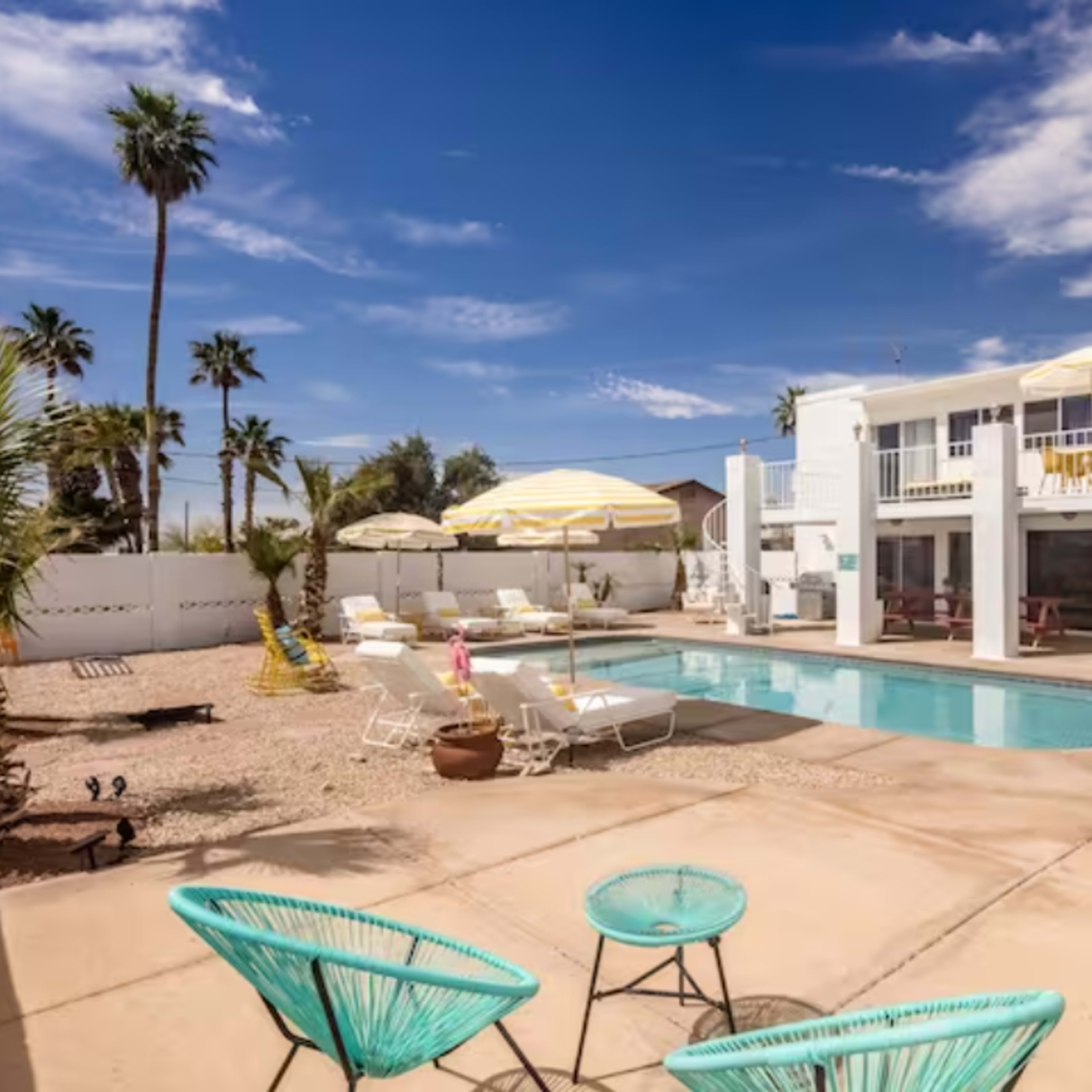 A sunny pool area surrounded by palm trees and lounge chairs, with a two-story white building in the background.