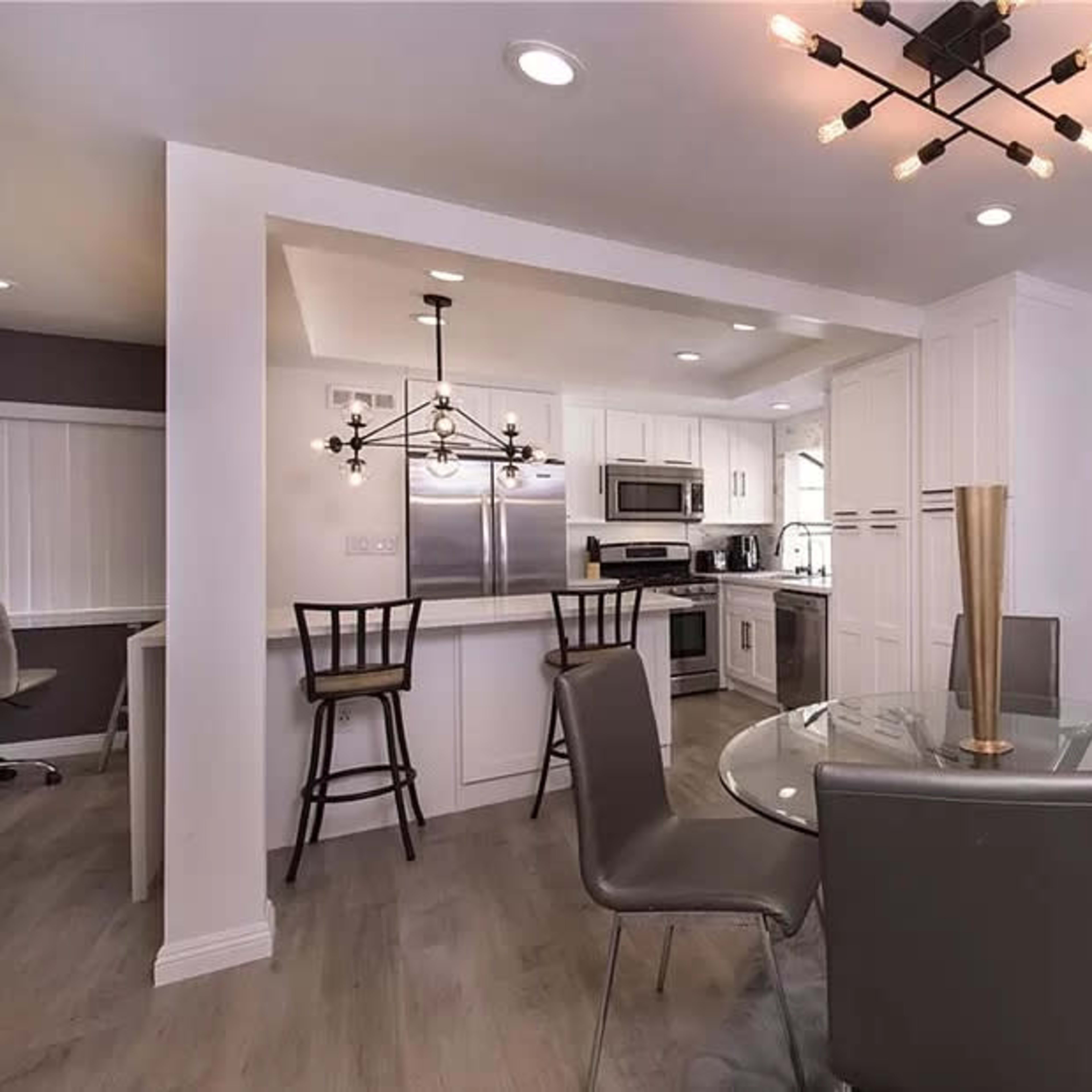A modern dining area features a round glass table surrounded by four gray chairs, with a kitchen visible in the background.
