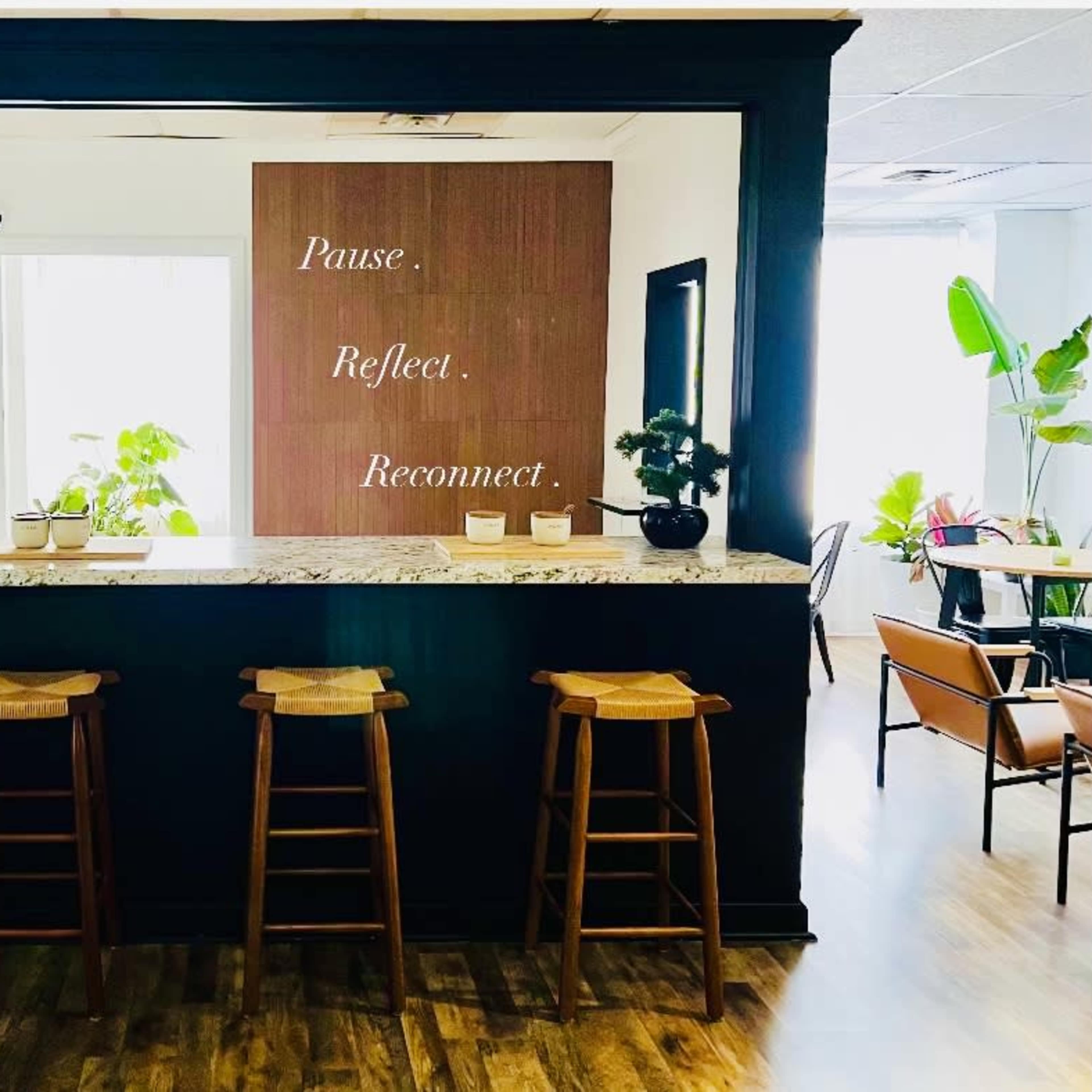 A modern reception area features a granite countertop with three wooden stools and a wall displaying the words "Pause. Reflect. Reconnect."