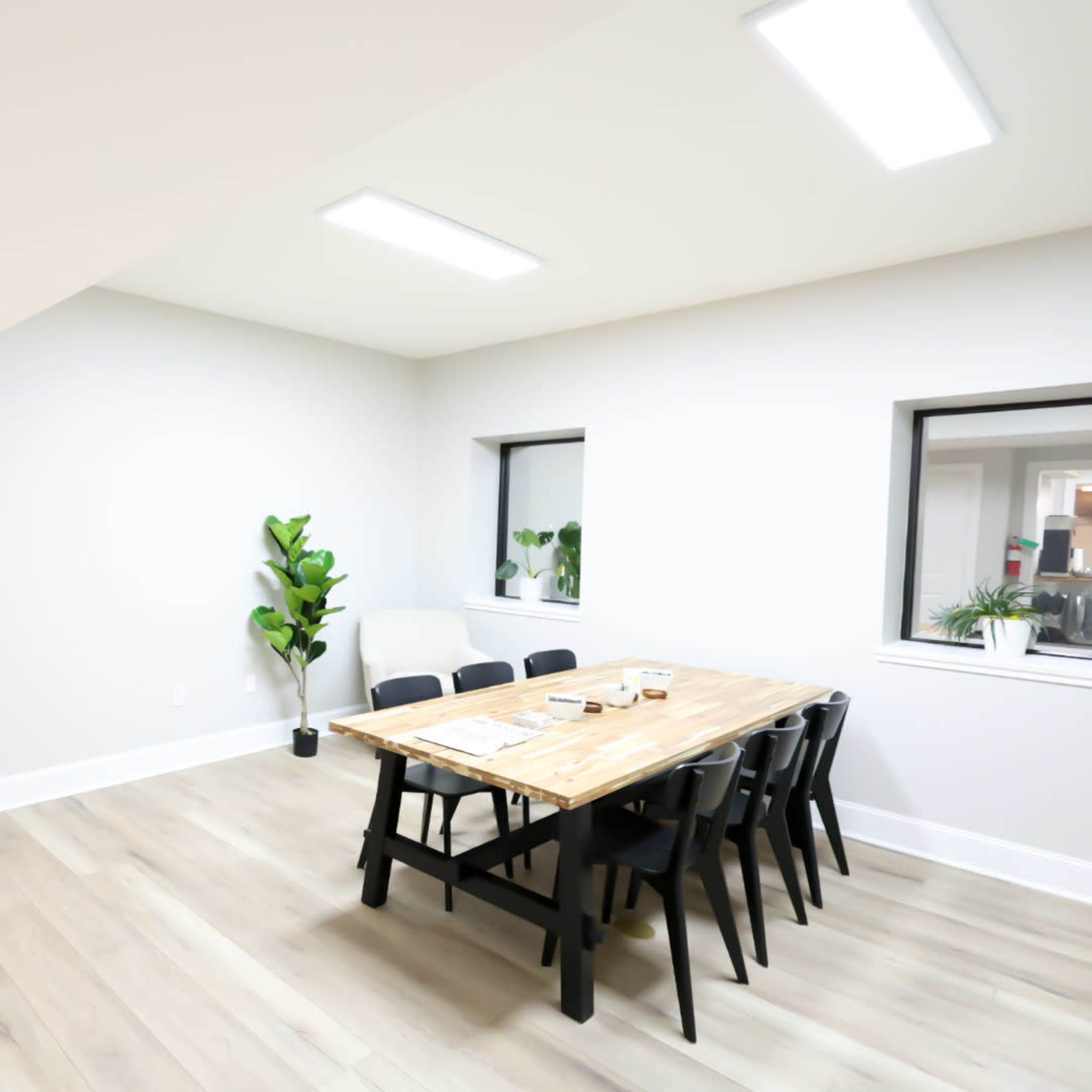 A well-lit, modern office space features a wooden table surrounded by black chairs, with plants and windows along the walls.