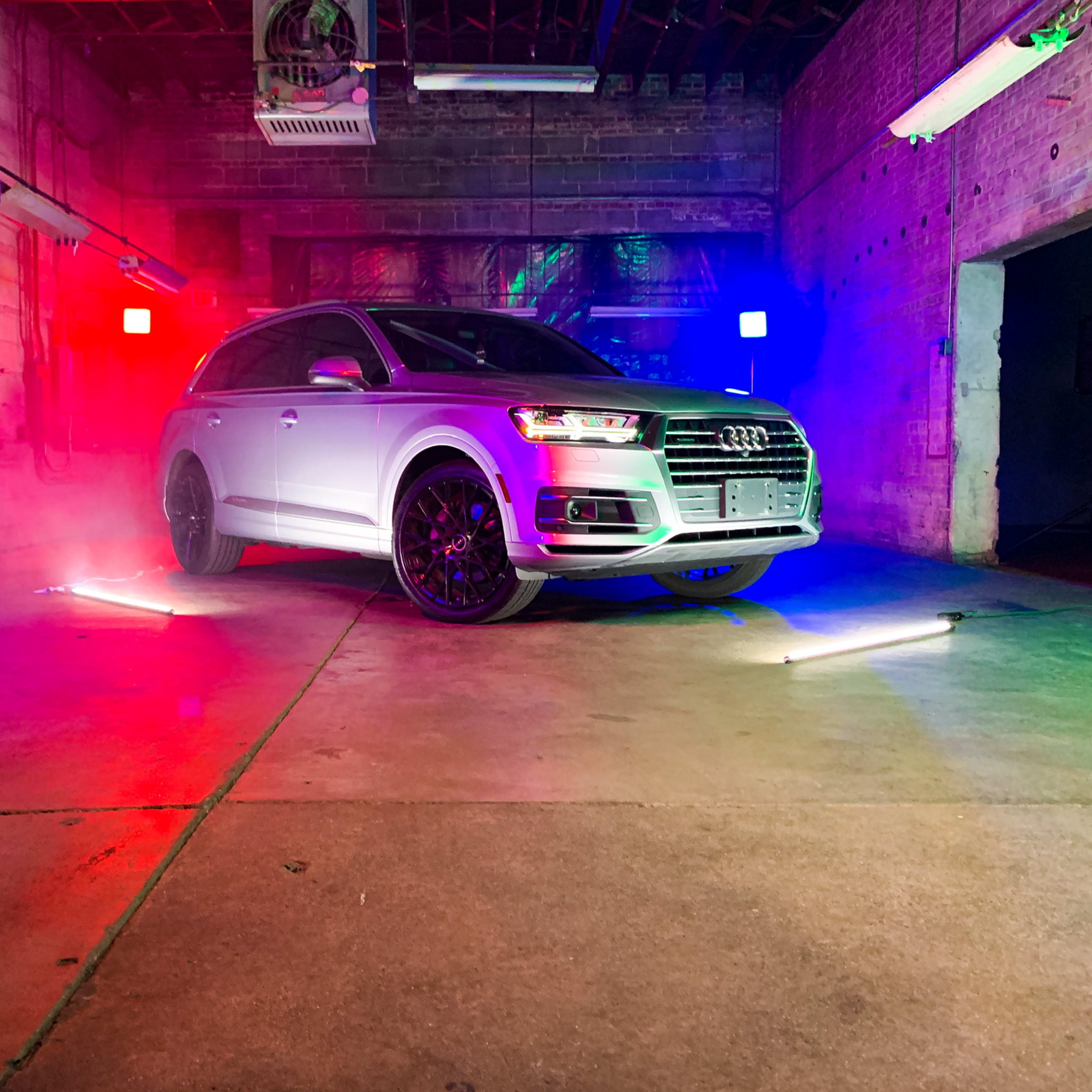 A silver Audi Q7 is positioned in a dimly lit industrial space, illuminated by red, blue, and green lights.