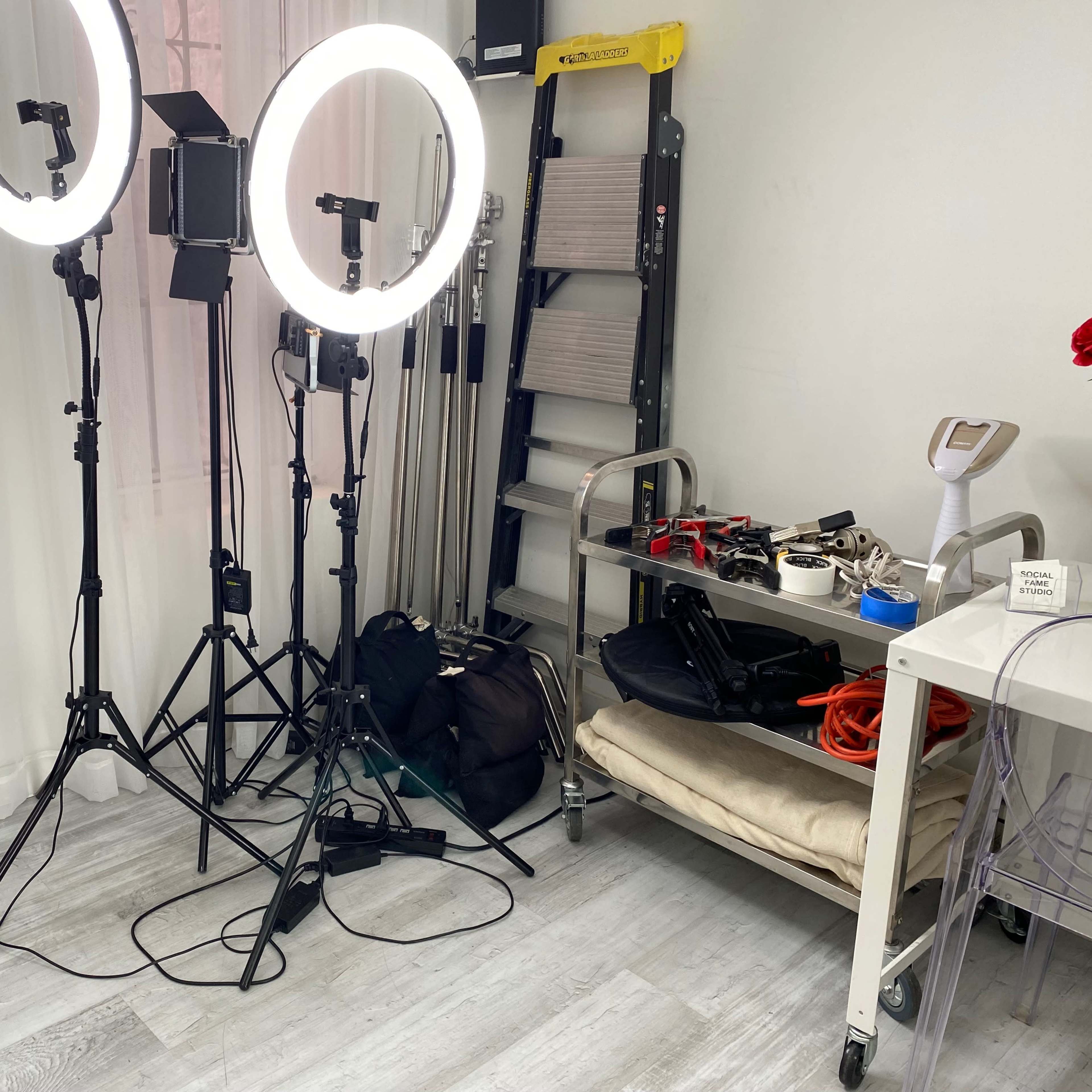 A photography setup with two ring lights, a stool, and various equipment on a table in a room with light-colored walls and flooring.