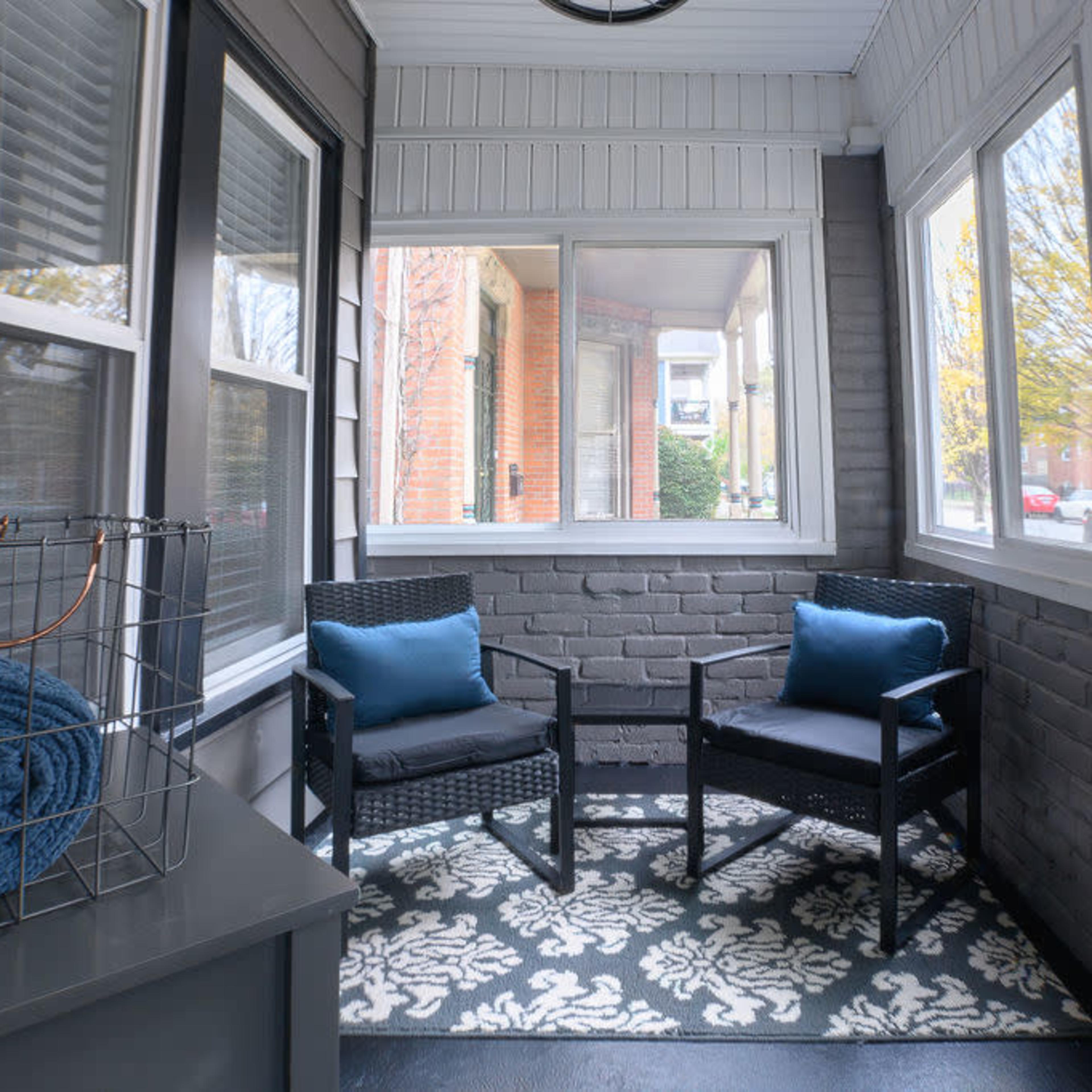 A cozy sunroom featuring two black chairs with blue cushions, a decorative rug, and large windows allowing natural light in.