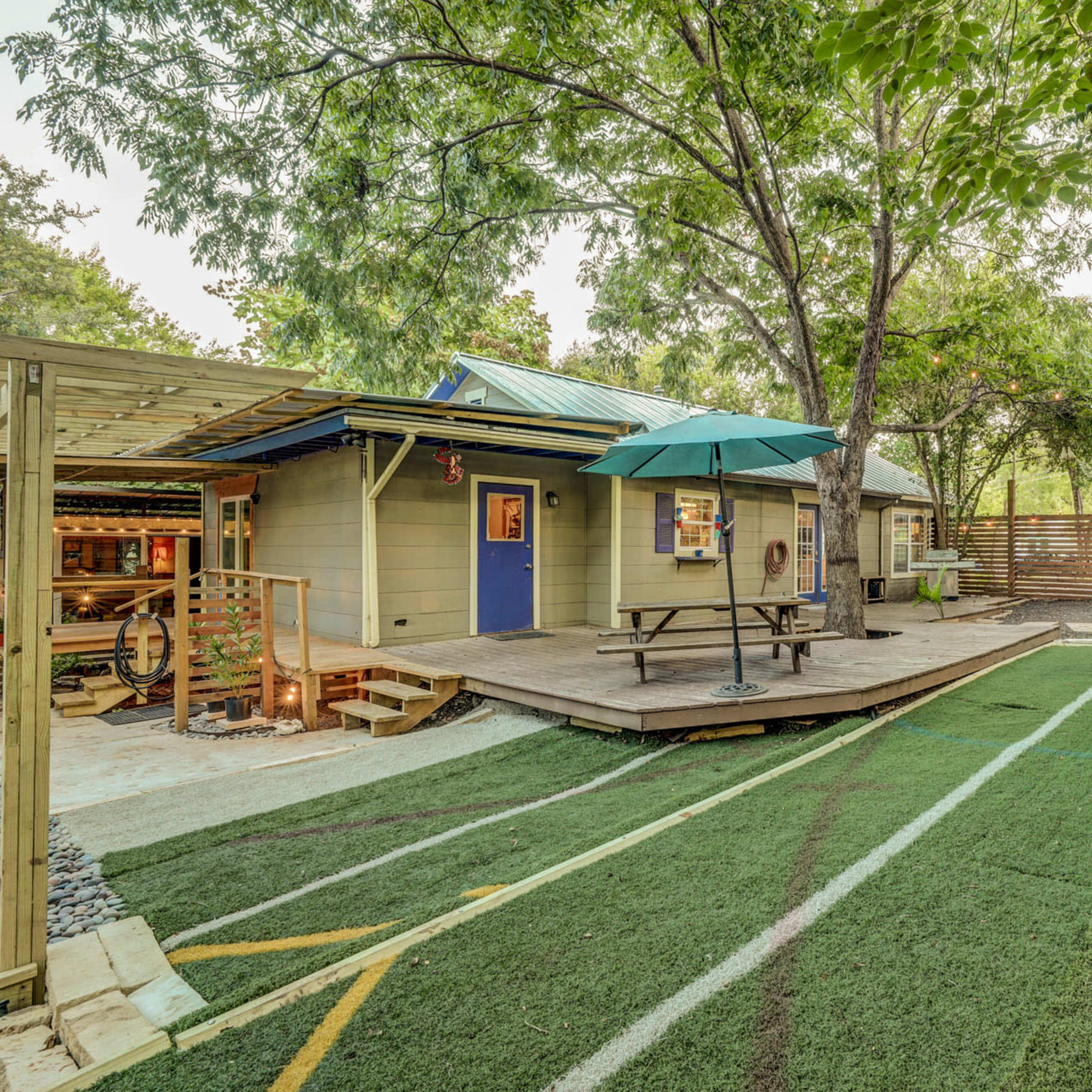 A landscaped backyard with artificial turf, a small deck with a table and umbrella, and a cozy seating area surrounded by wooden fencing and trees.