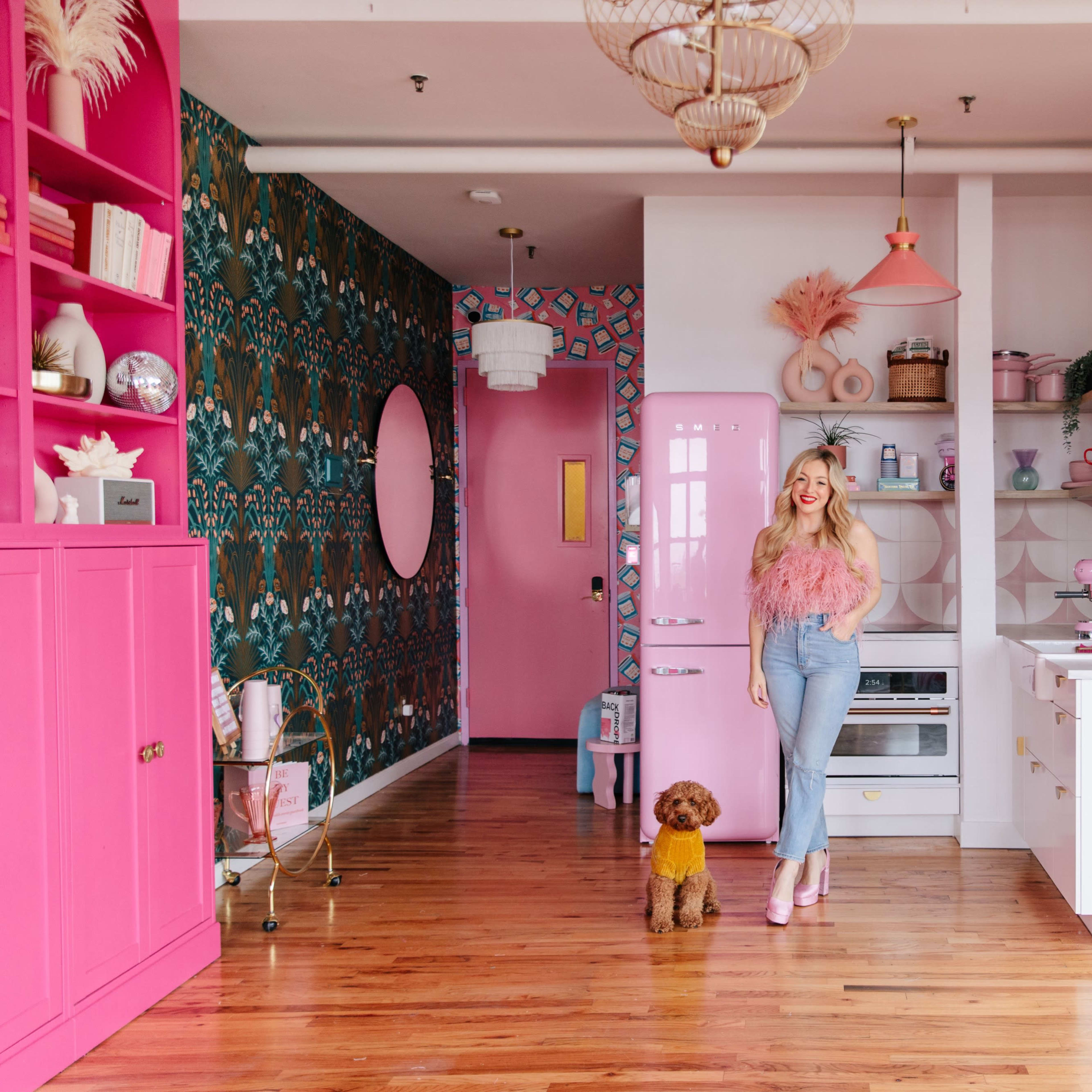 A woman stands in a brightly decorated kitchen with pink accents and floral wallpaper, next to a small brown dog.