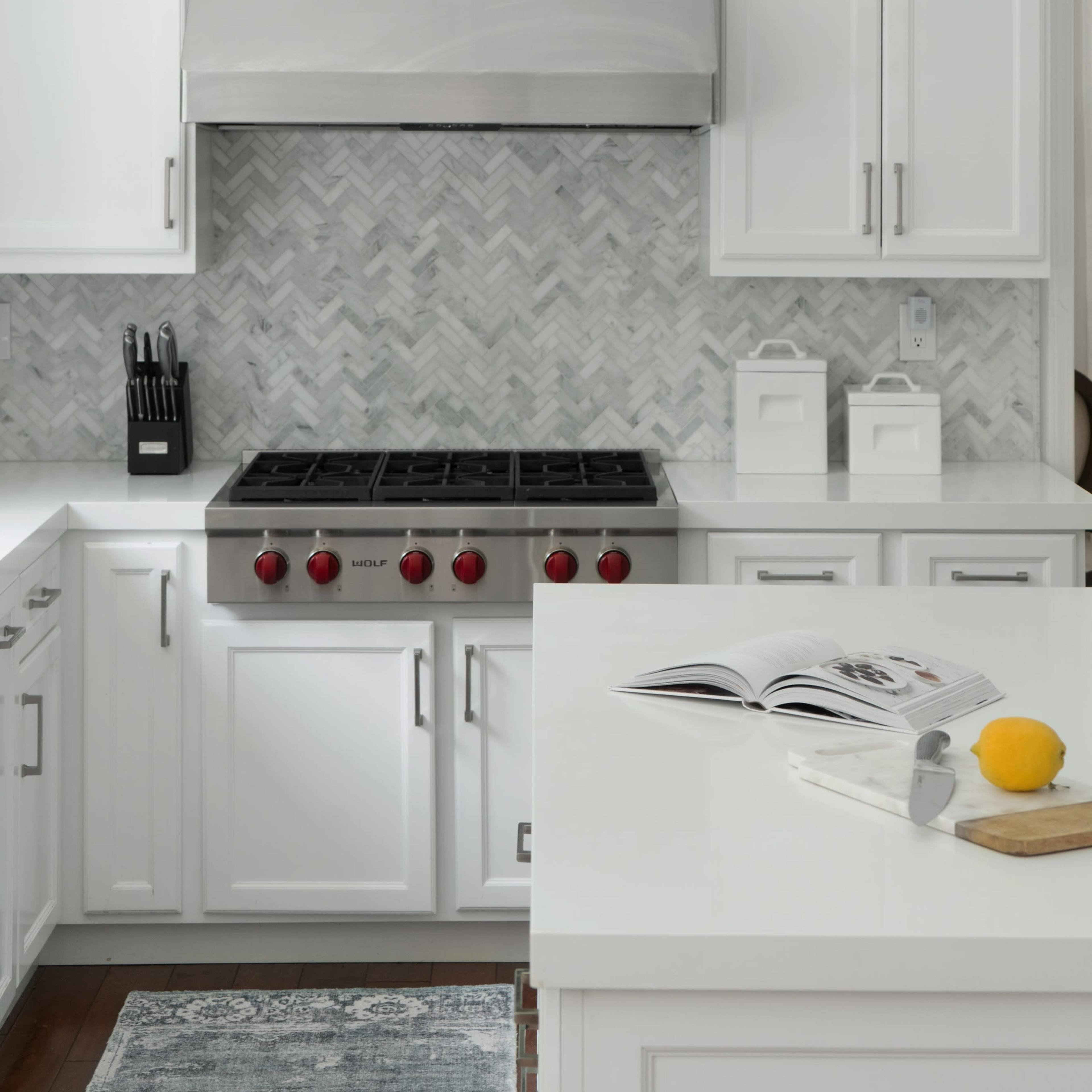 A modern kitchen features white cabinetry, a stainless steel oven hood, and a herringbone-patterned backsplash.