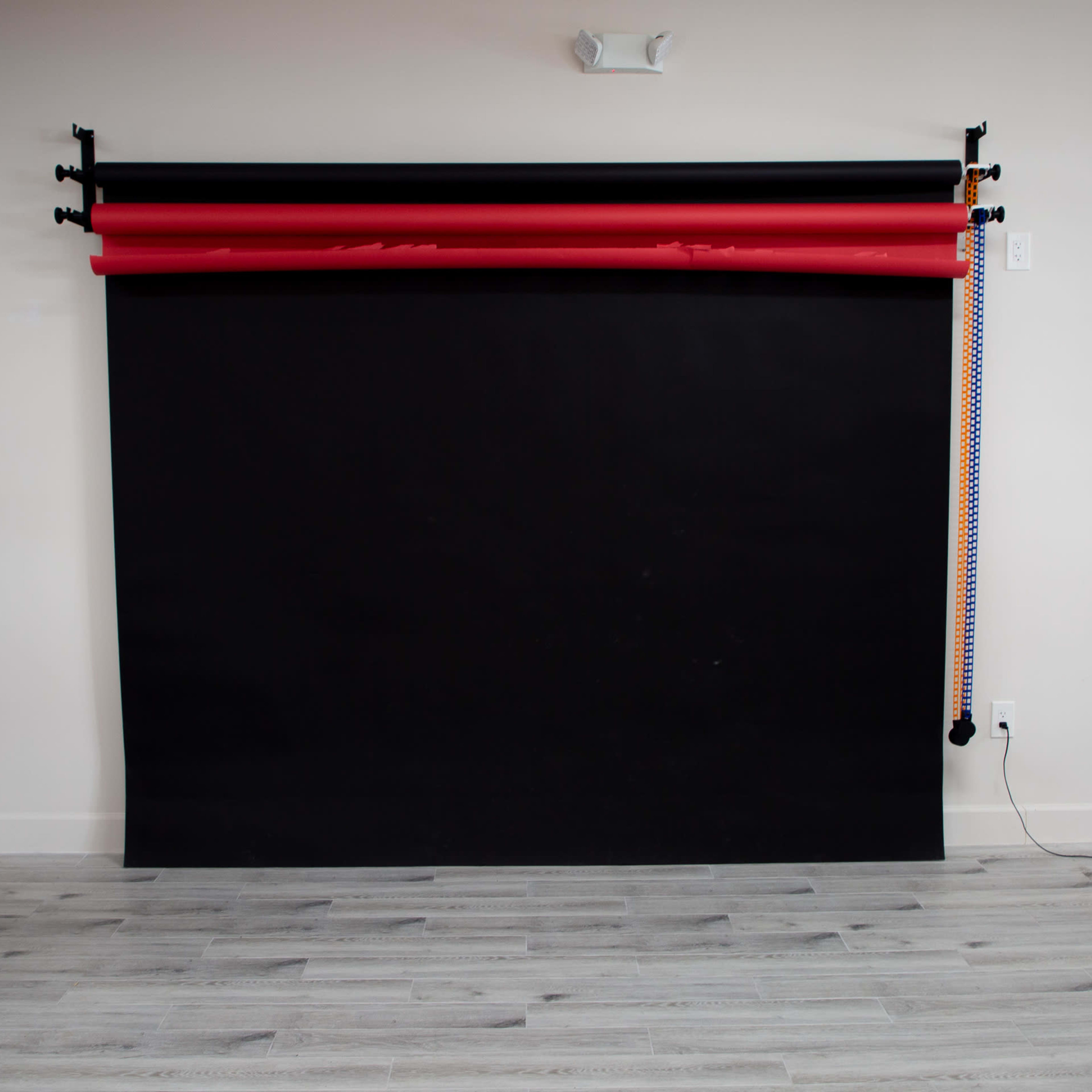 A dark backdrop setup with red and orange studio rolls suspended from a bar on a wall, with wooden flooring visible below.