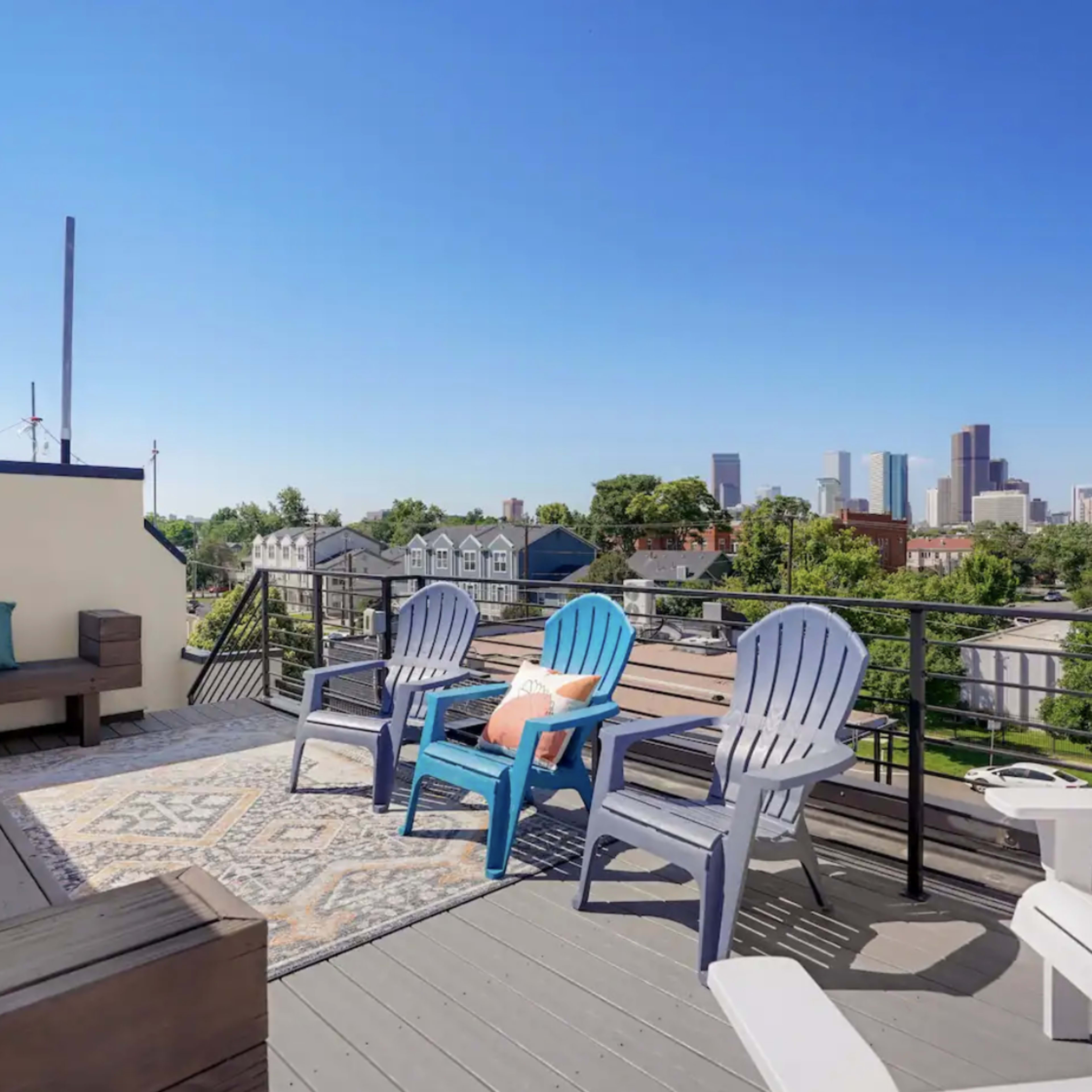 A rooftop terrace features blue and white Adirondack chairs on a wooden deck, overlooking a city skyline with clear skies.
