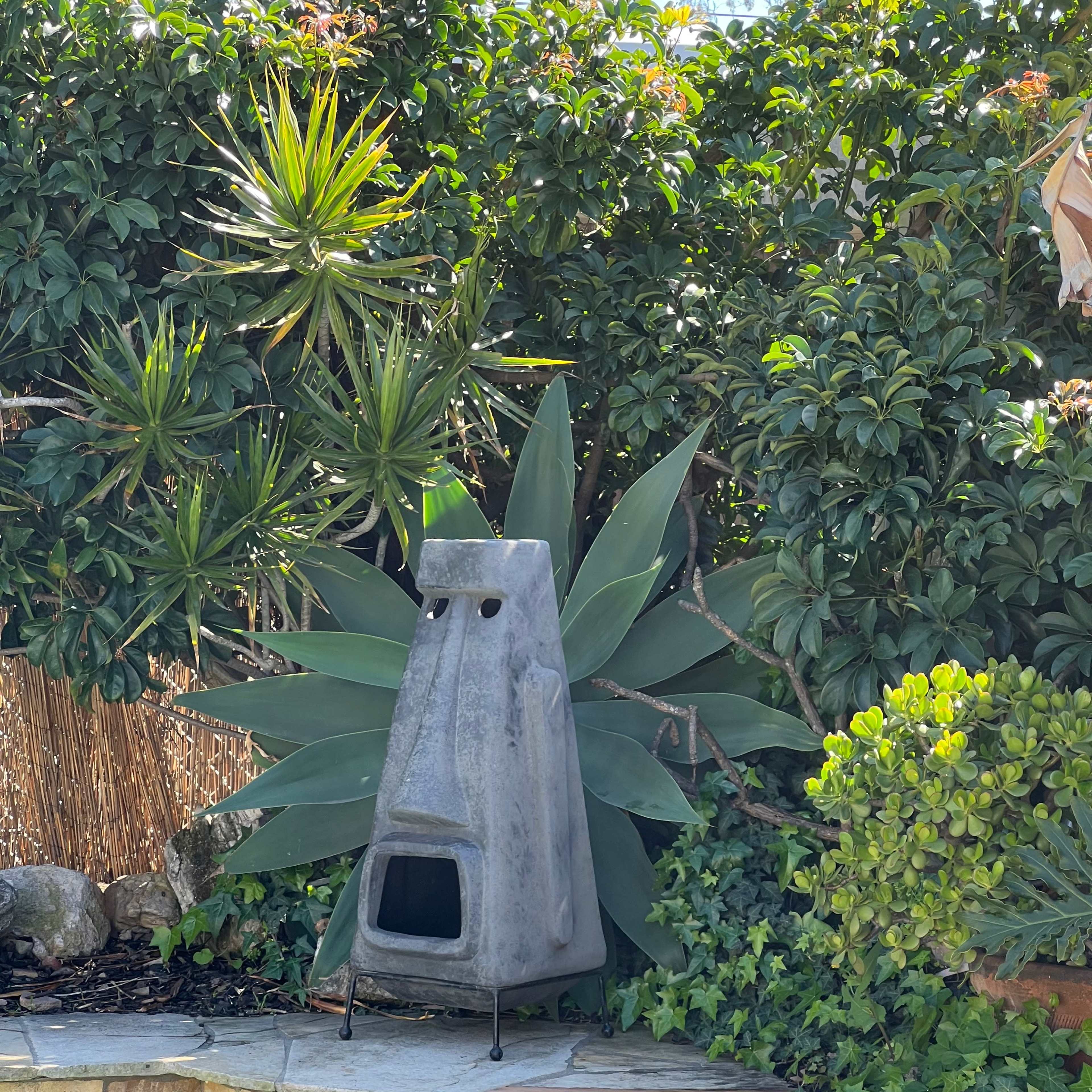A concrete outdoor fireplace stands near a swimming pool, surrounded by lush greenery and plants.