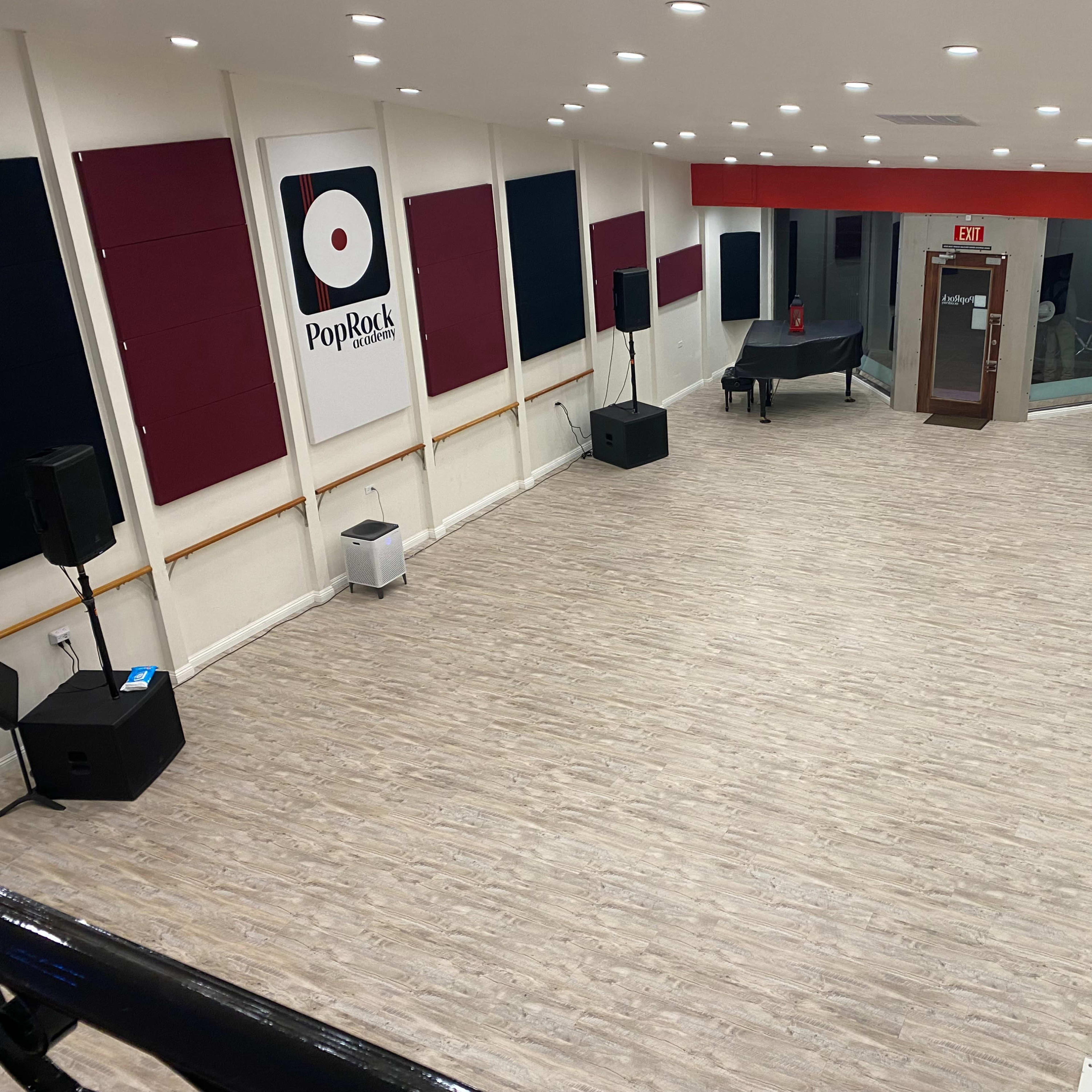 A spacious interior room with a wooden floor, featuring sound equipment, a grand piano, and red and black wall panels, along with a glass entrance.